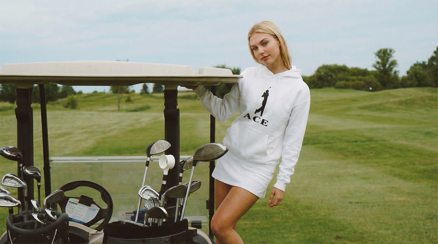 A young woman in a white hoodie standing next to a golf cart filled with golf clubs on a golf course.