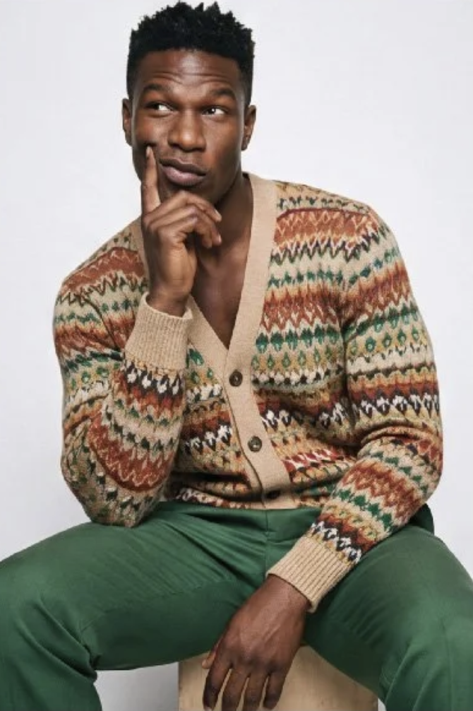 A photo of a man with short curly hair, wearing a colorful patterned cardigan and green pants, sitting against a plain white background. He has a thoughtful expression with his index finger resting on his cheek.