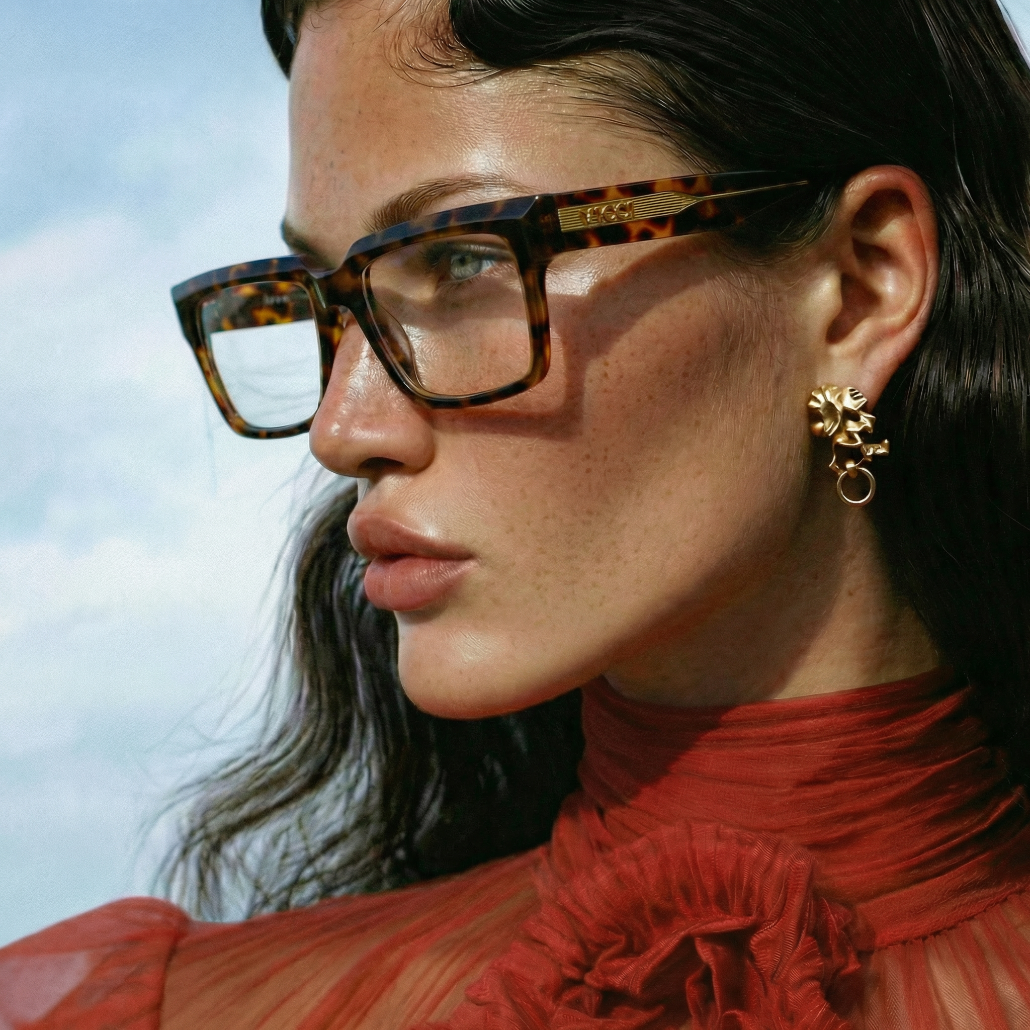 A close-up photo of a woman's face with black hair, wearing tortoiseshell eyeglasses with gold detailing, gold earrings, and a red patterned top.