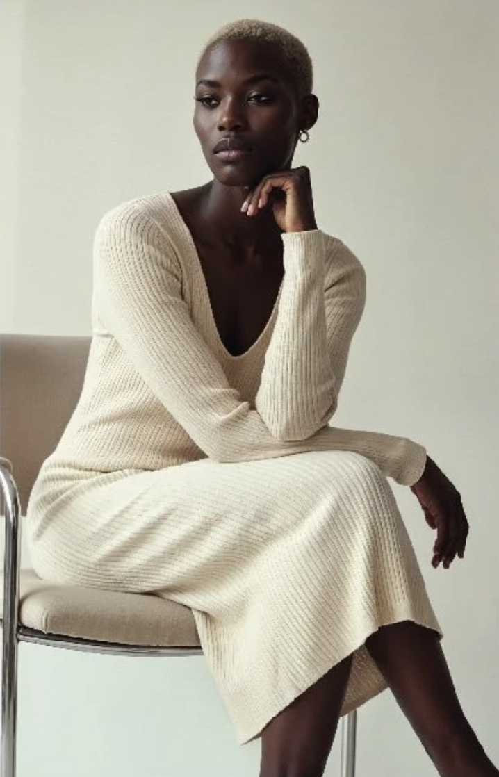 A woman with dark skin and short blonde hair sits on a beige chair, wearing a cream-colored knit dress, posing with her chin resting on her hand against a plain background.