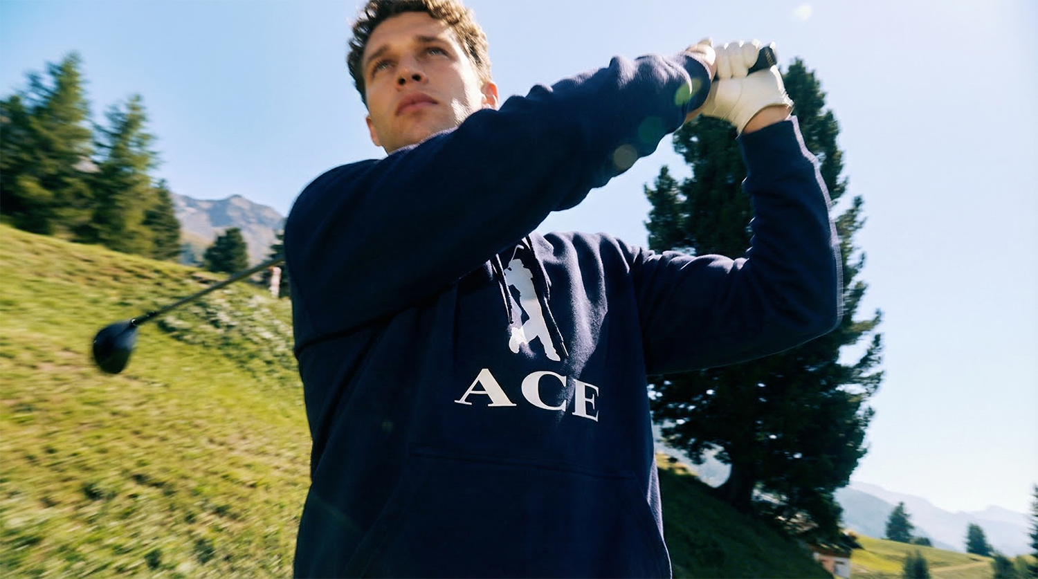 Young man in a navy hoodie swinging a golf club outdoors in a scenic landscape with trees and mountains in the background.