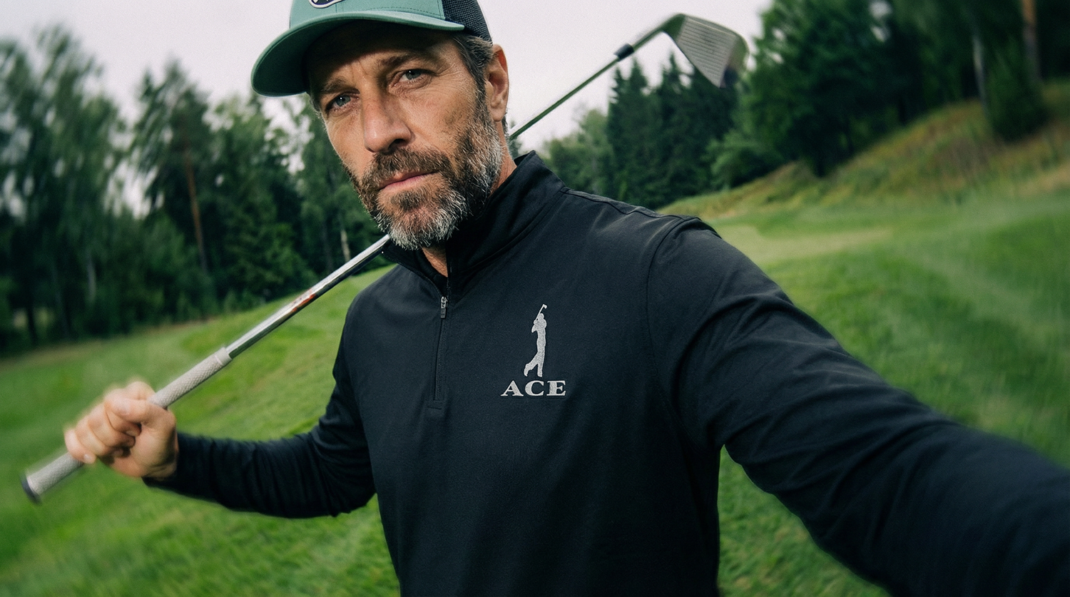 A man with a beard wearing a cap and black zip-up jacket holding a golf club on a golf course with green grass and trees in the background.