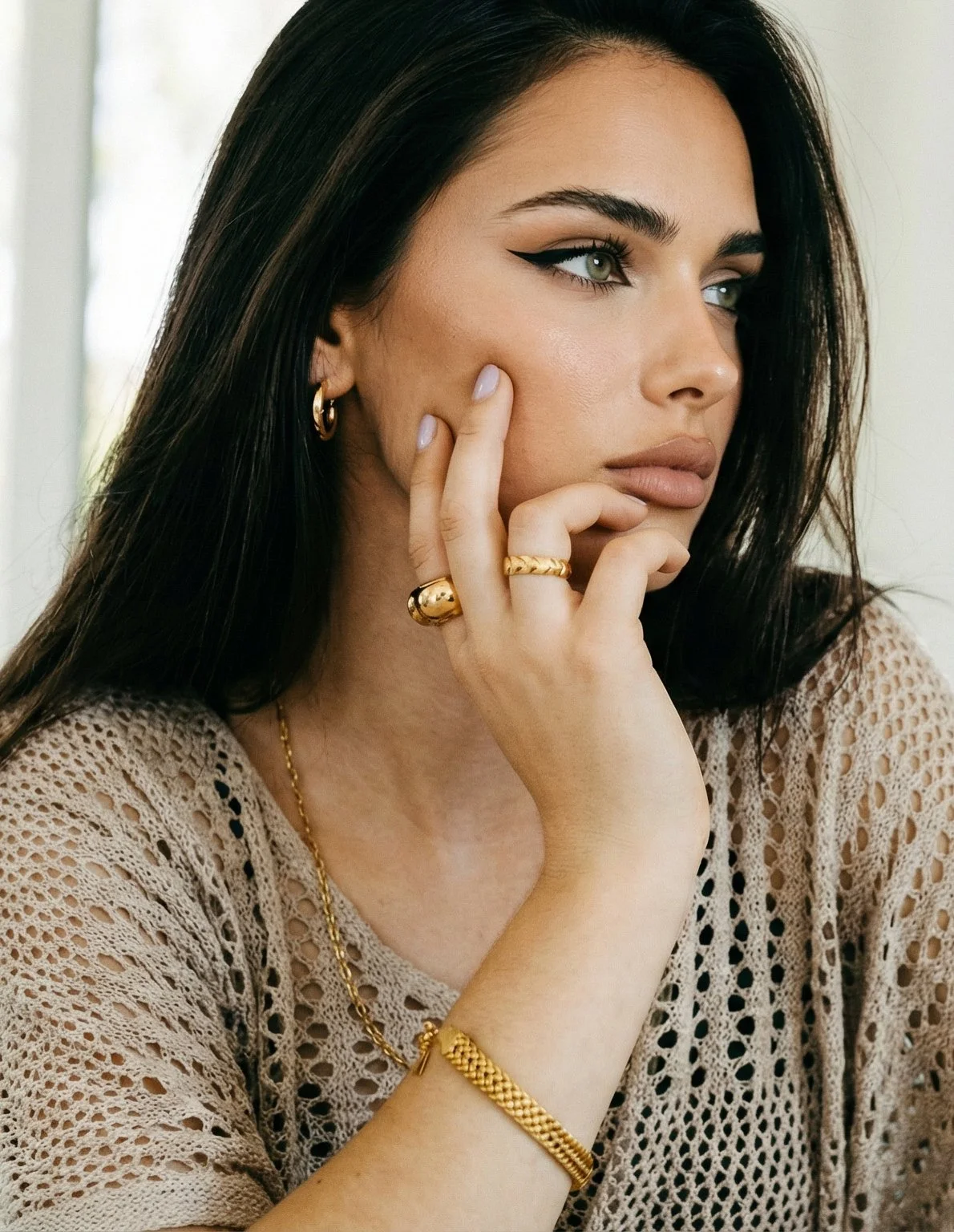 A woman with dark hair and light skin is wearing gold jewelry, including earrings, necklaces, rings, and a bracelet, and is dressed in a beige crocheted top. She is posing with her hand on her face, gazing thoughtfully to the side.