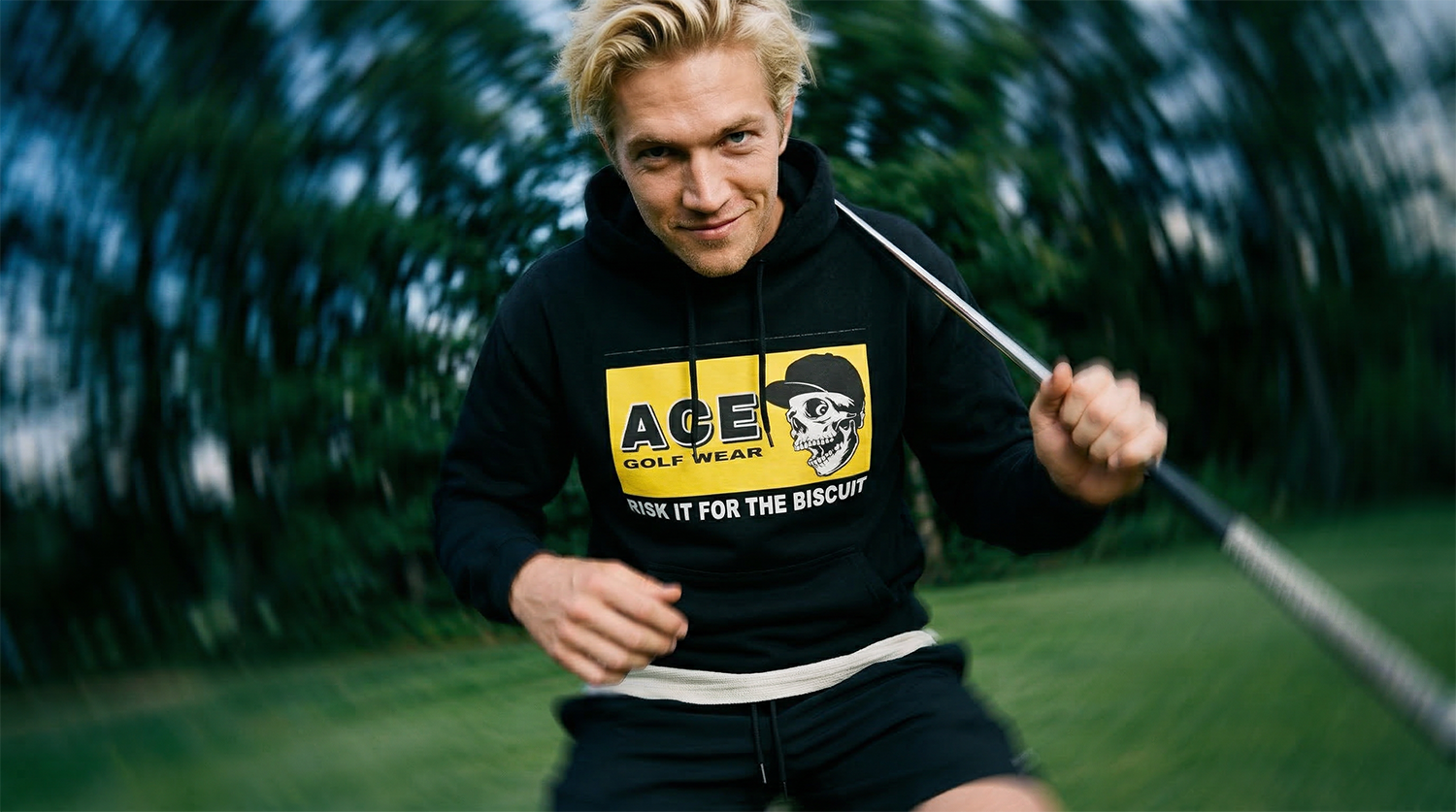 A young man with blond hair smiling while holding a golf club on a grassy golf course, wearing a black hoodie with a graphic and text.