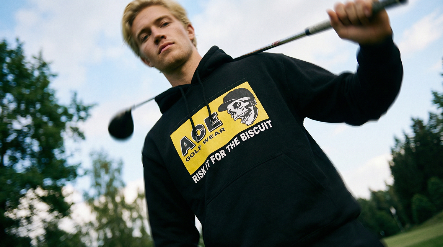 A young man in a black hoodie holding a golf club over his shoulder outdoors, with trees and a cloudy sky in the background.