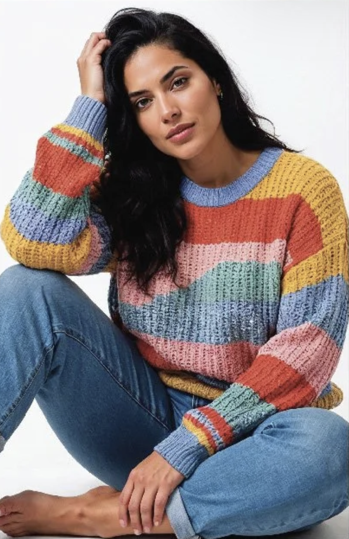 A woman with long black hair, wearing a colorful striped sweater and blue jeans, sitting on the floor with one leg bent and her hand resting on her head, looking at the camera.