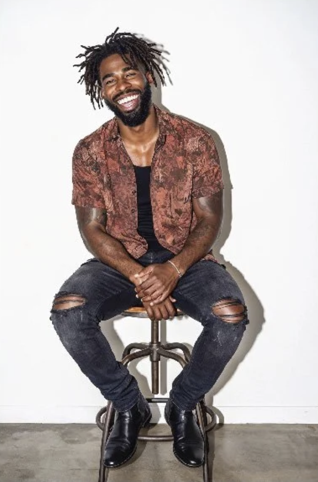 Man with dreadlocks and a beard, smiling and sitting on a stool against a plain white background, wearing a patterned shirt, ripped jeans, and black boots.