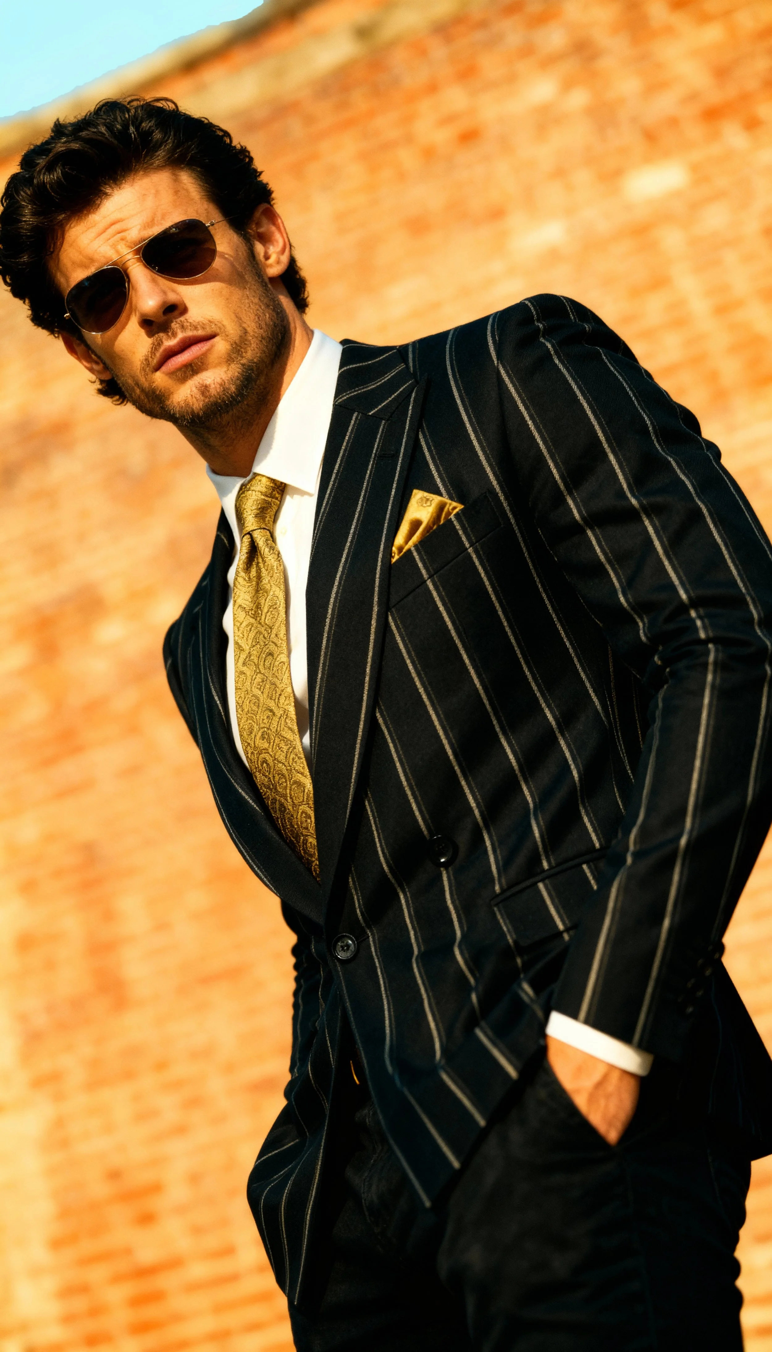 Stylish man wearing sunglasses, a dark pinstripe suit, a white shirt, and a gold tie, standing against a blurred brick wall background.