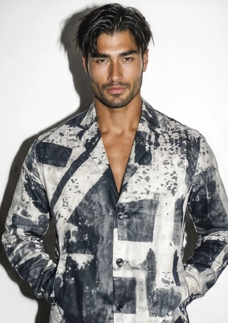 A man with dark, wavy hair and light facial hair, wearing a black and white patterned button-up shirt, standing against a white background.