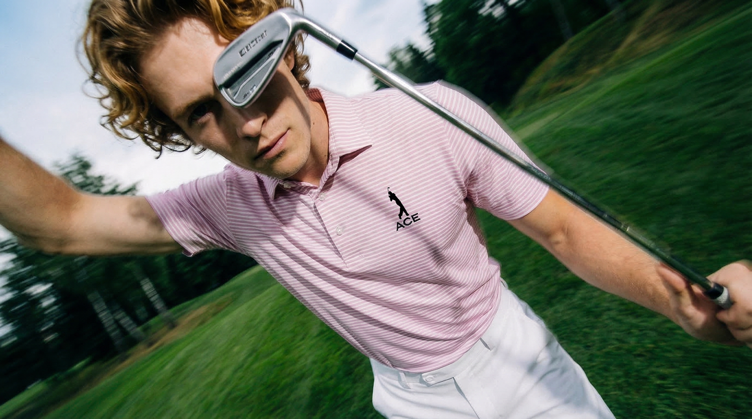 A man with curly red hair wearing a pink striped polo shirt with the logo 'ACE' and white pants, holding a golf club on a golf course.