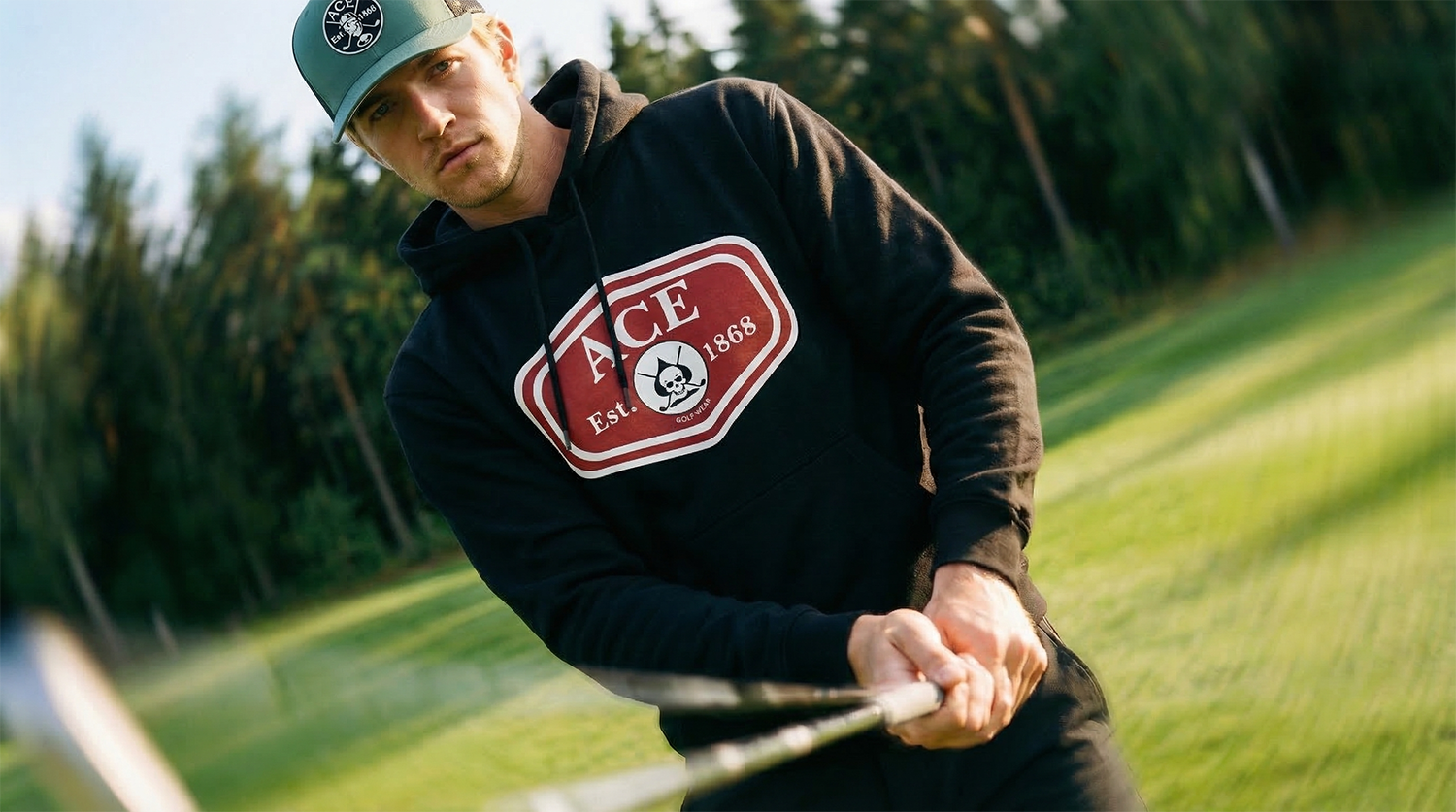 A man playing golf on a golf course during the daytime, holding a golf club and focusing on his shot. He is wearing a hoodie and a gray cap with the ACE logo.