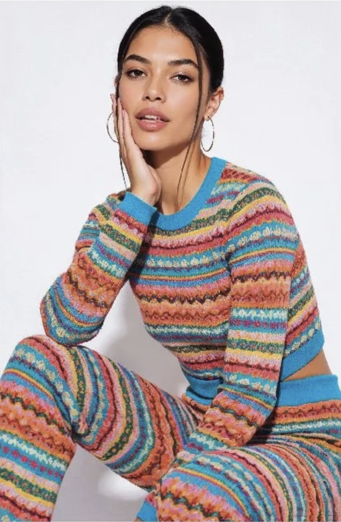 A young woman with long dark hair, wearing large hoop earrings, touches her face with her left hand. She is dressed in a colorful, patterned knit sweater and matching pants, sitting against a plain white background.