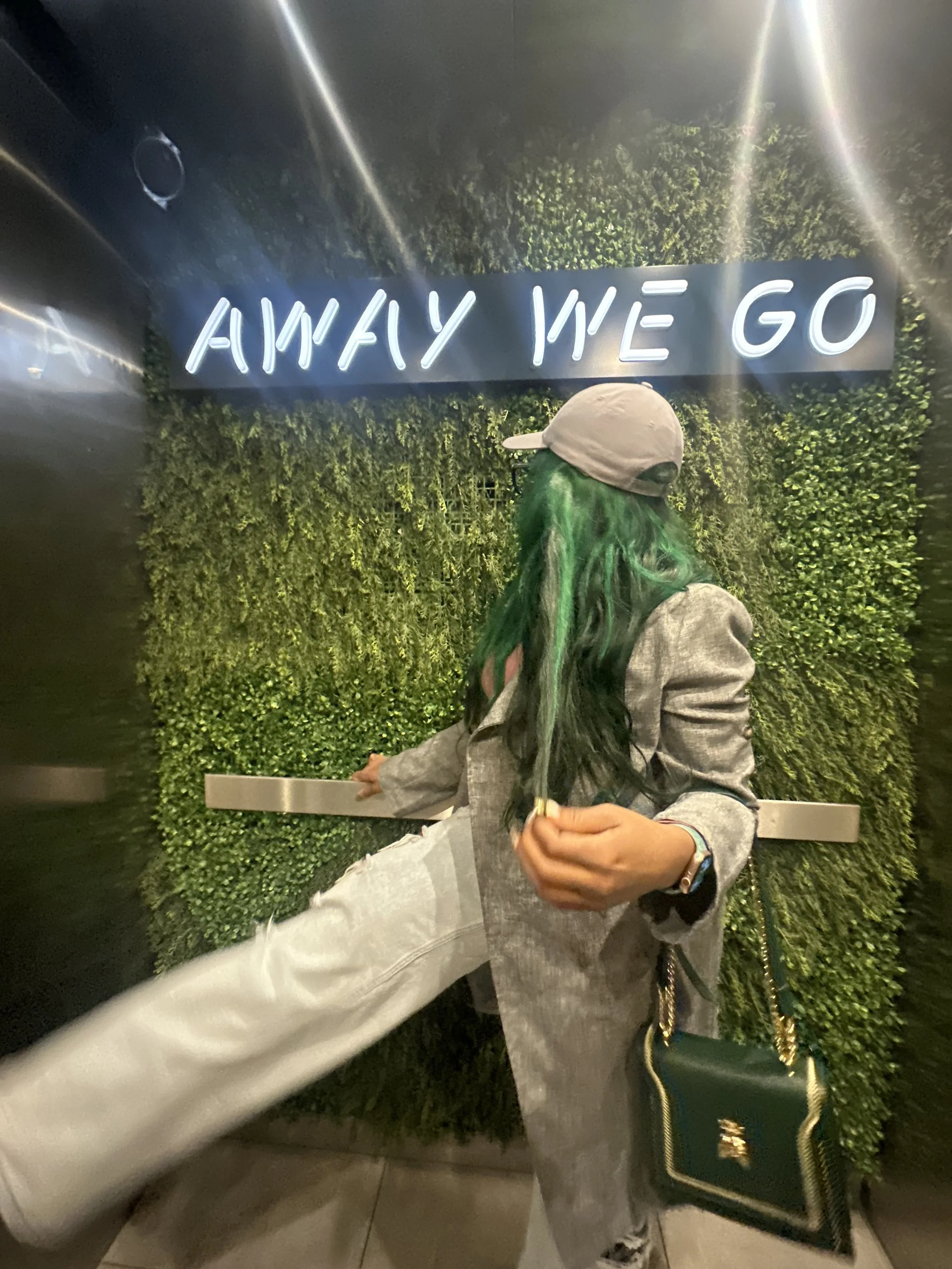 Person in green coat and cap standing near a wall with greenery, holding a handbag, beneath a neon sign reading 'AWAY WE GO'.
