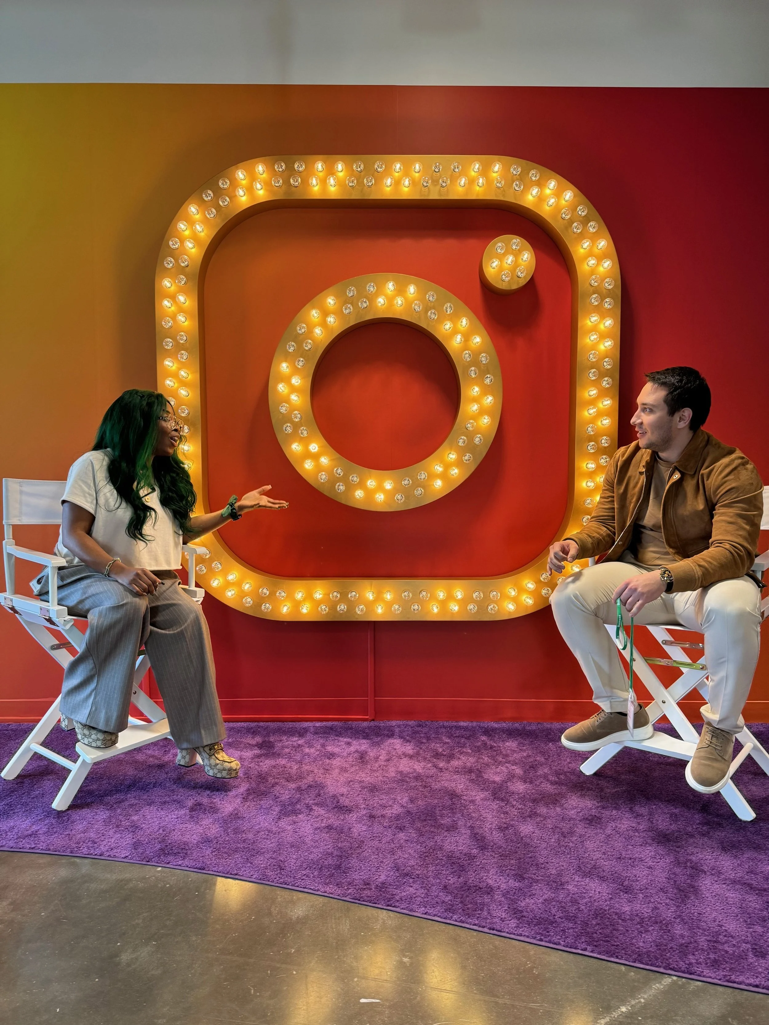 Two people sitting and talking in front of a large illuminated Instagram logo, with vibrant red and orange background and purple carpet.