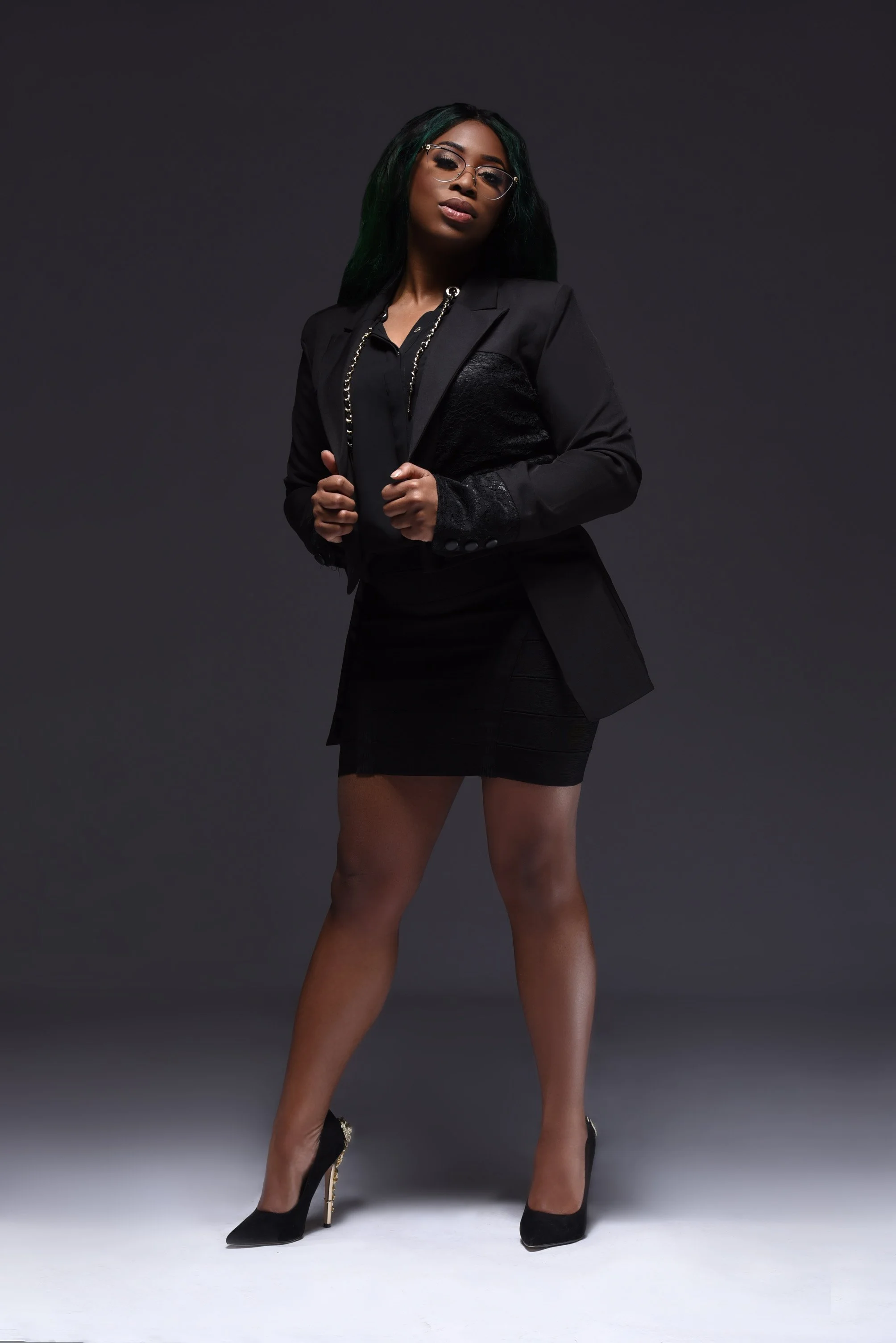 A woman with long dark green hair and glasses in a black outfit, posing confidently against a dark background.