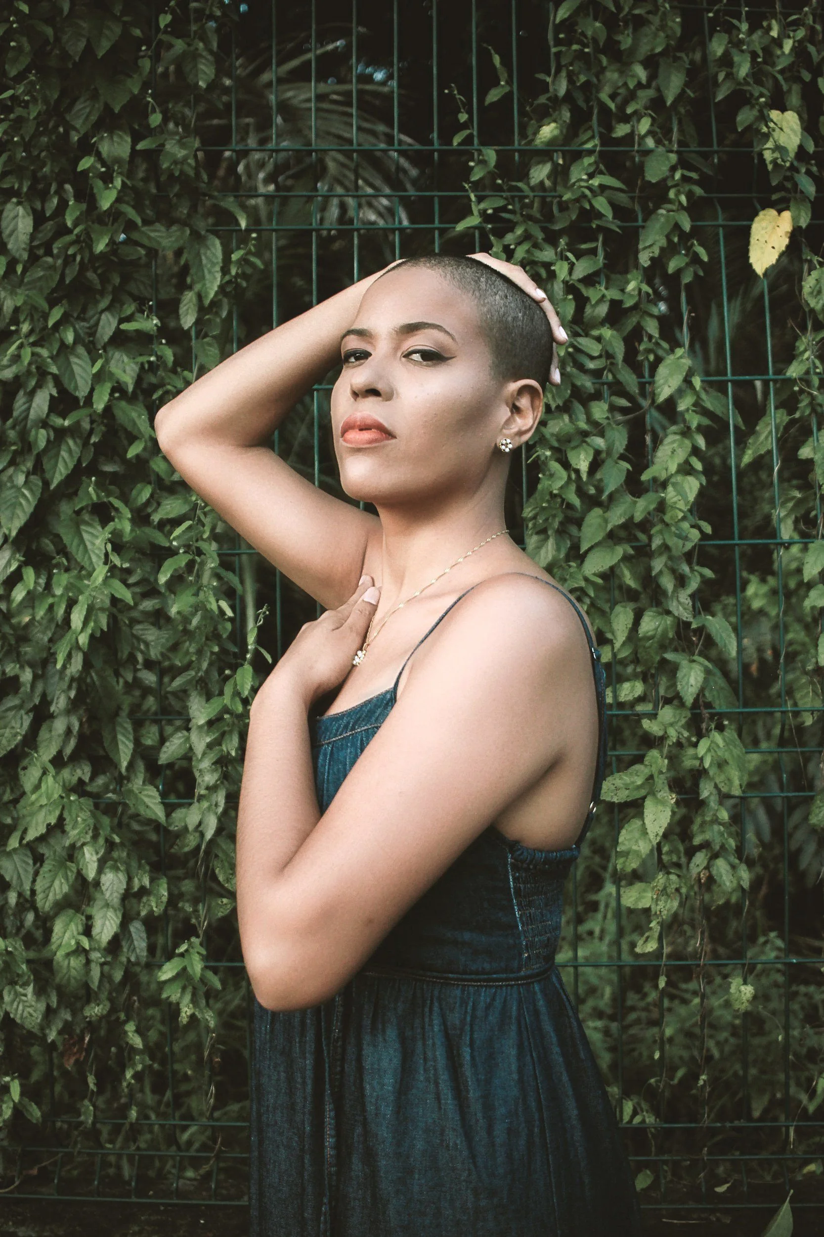 A woman with a shaved head wearing a dark denim sleeveless dress standing in front of a green leafy background and a metal fence. She has her right hand on her head and her left hand on her chest, looking confidently at the camera.