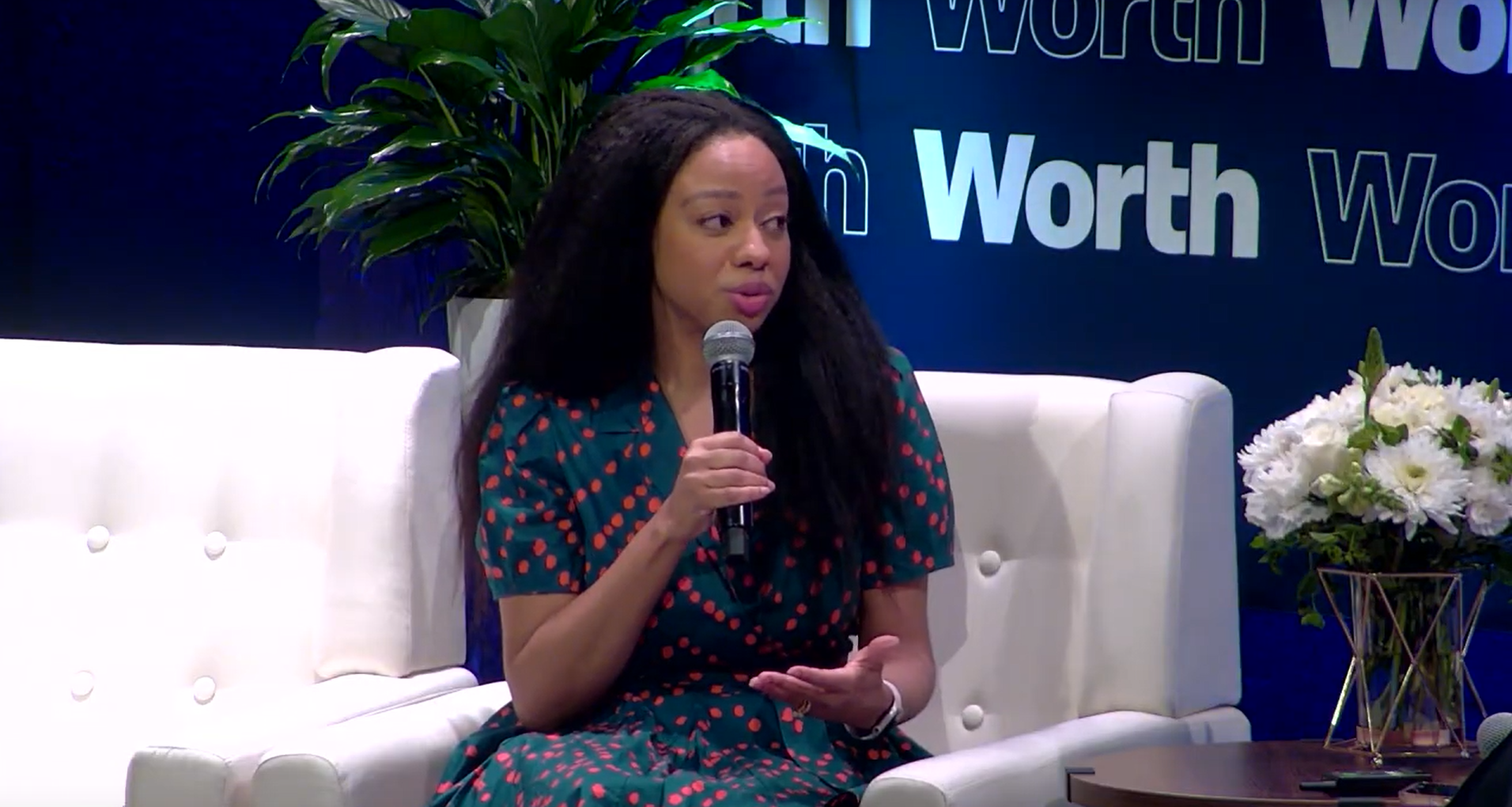 A woman with long curly black hair speaking into a microphone while seated on a white couch. She is wearing a green dress with red polka dots. Behind her is a large blue background with the word 'Worth' repeatedly written. There are green plants and a vase with white flowers on a table next to her.