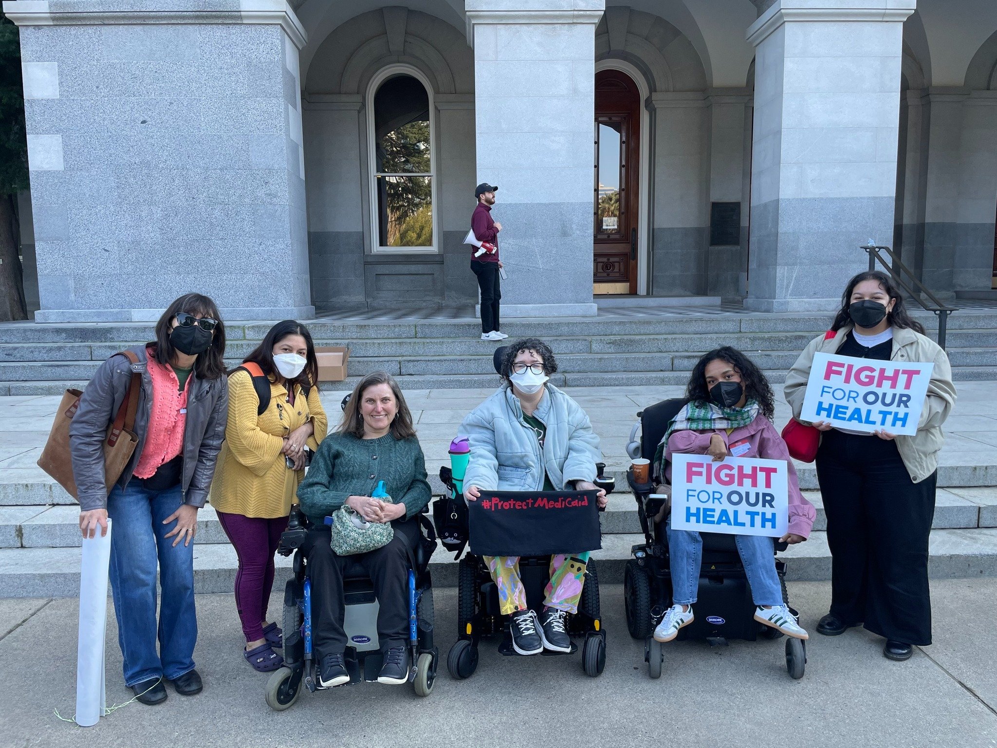 Un grupo multirracial de seis personas, tres en sillas de ruedas, la mayoría con máscaras, sosteniendo carteles frente al edificio del capitolio estatal que dicen "Lucha por nuestra salud" y "#ProtectMedicaid".