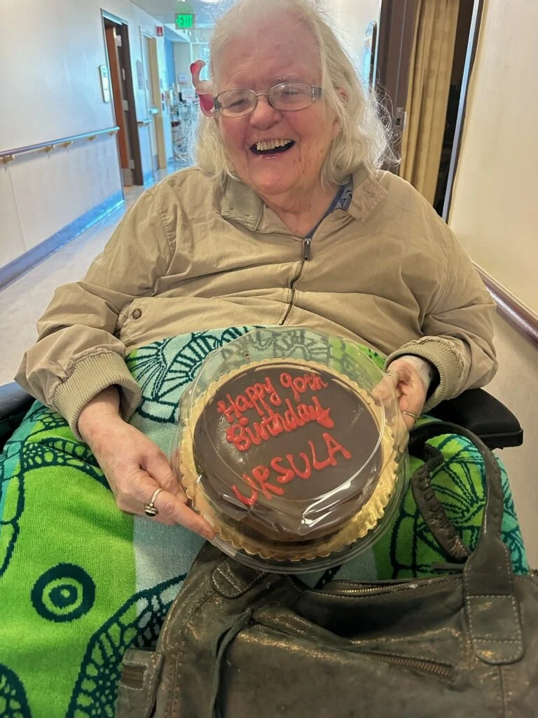 A smiling woman with white hair sitting in a wheelchair, holding a cake that says "Happy 90th birthday Ursula"