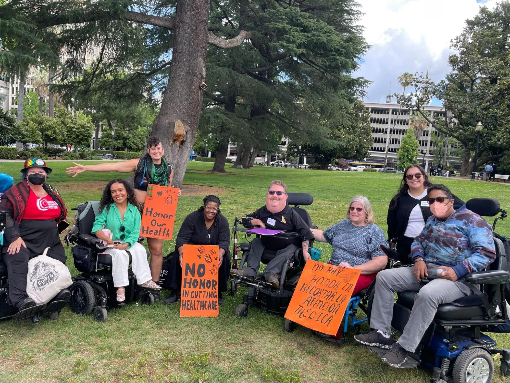 A multiracial group of eight people, many of them in wheelchairs and some wearing masks, outside wearing signs that say "honor our health" and "no honor in cutting healthcare"