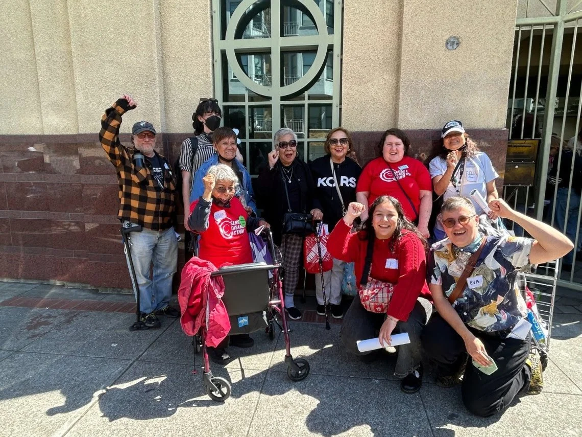 A group of ten people outside in front of a stone building, some standing or squatting, some using mobility aids. Many are smiling with fists in the air.