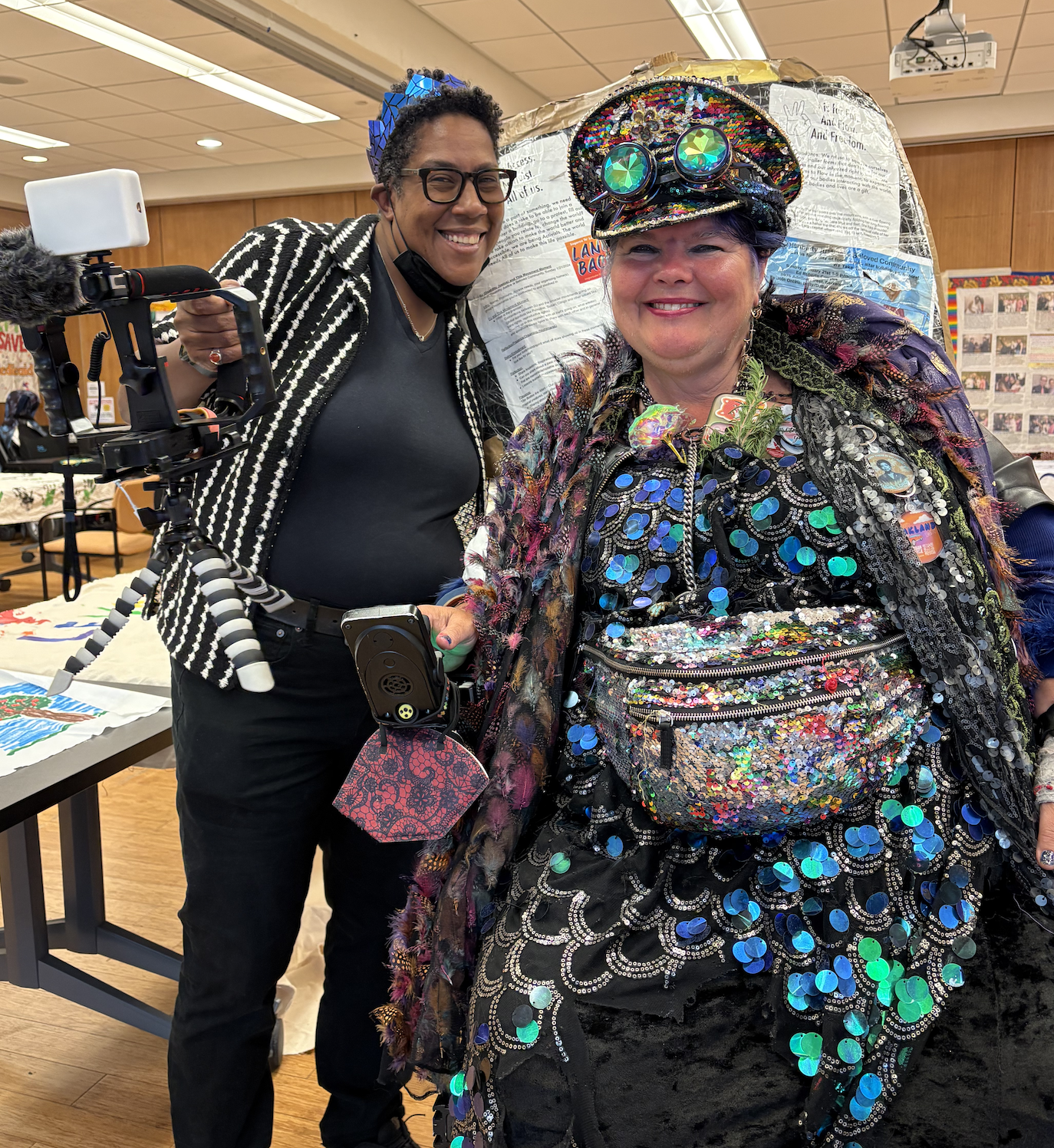 Two people smiling, one standing and holding a camera and microphone, one in a wheelchair. Both are wearing sparkly headwear and one of them is in a sequined outfit with a sequined bag.