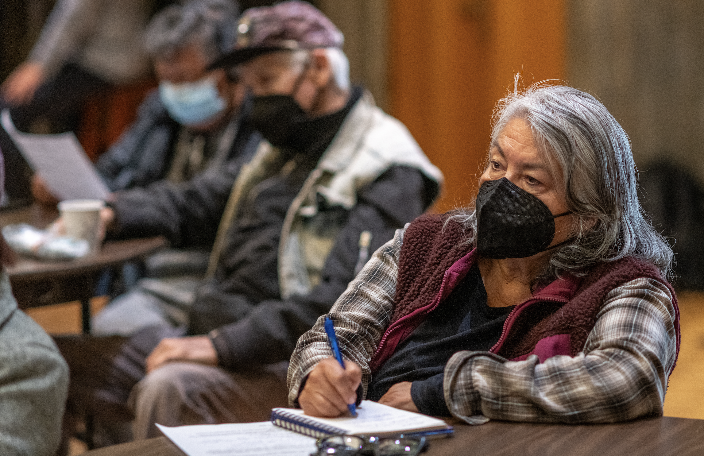 A woman masked woman sits at a table writing on a piece of paper, with other masked people sitting behind her in the background