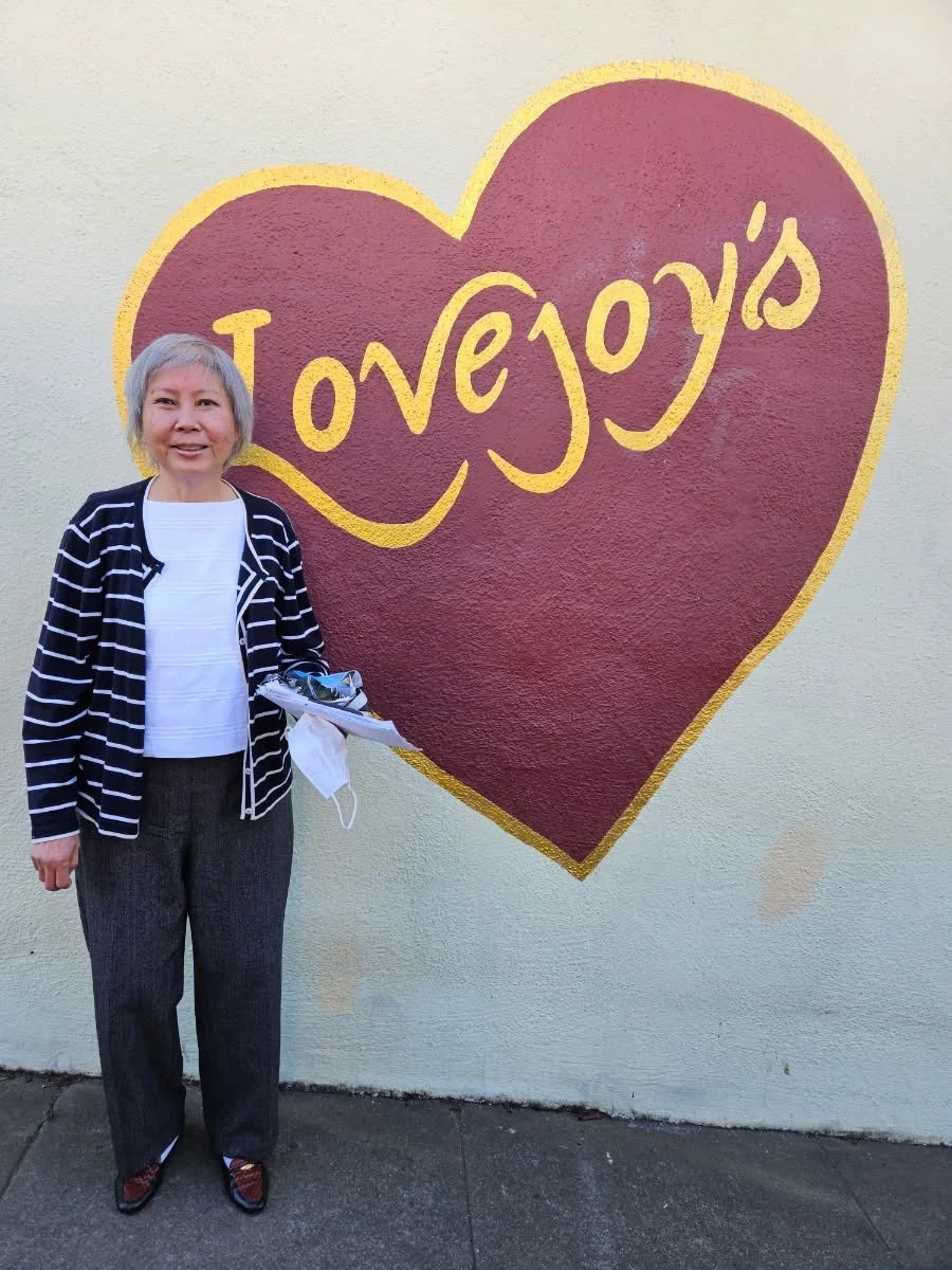A woman with short gray hair stands in front of a sign in the shape of a heart that says Lovejoy's