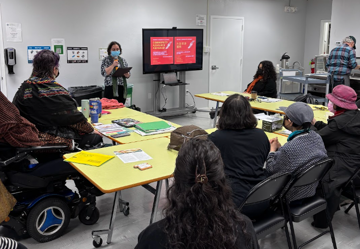 A group of people sits at a table in a horseshoe shape. At the front of the room is a woman standing next to a monitor with a presentation on it. Everyone is masked.