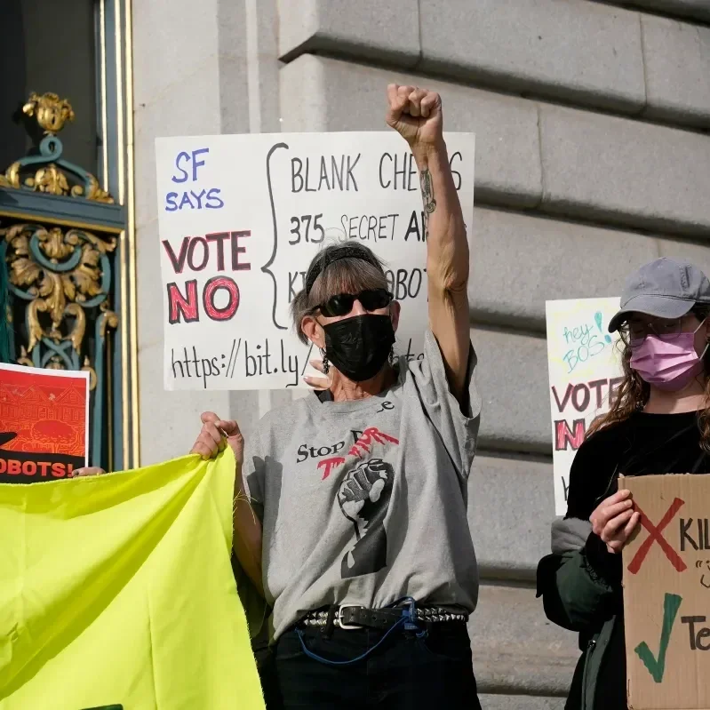 A woman with gray hair and a black mask standing with her first in the air in front of a sign that says "SF says vote no"