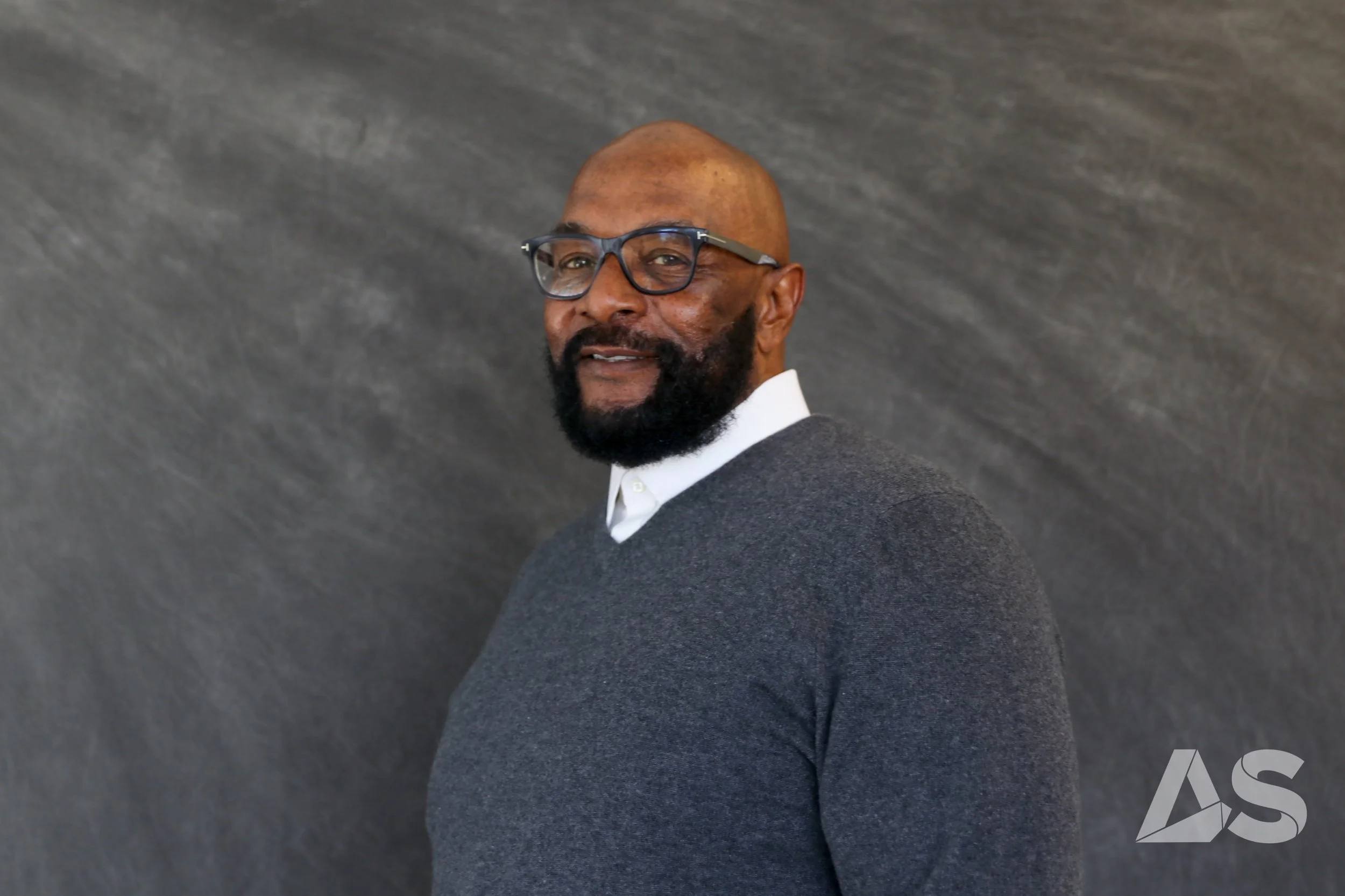 A man with a beard and glasses smiling, wearing a sweater with a collared shirt under it