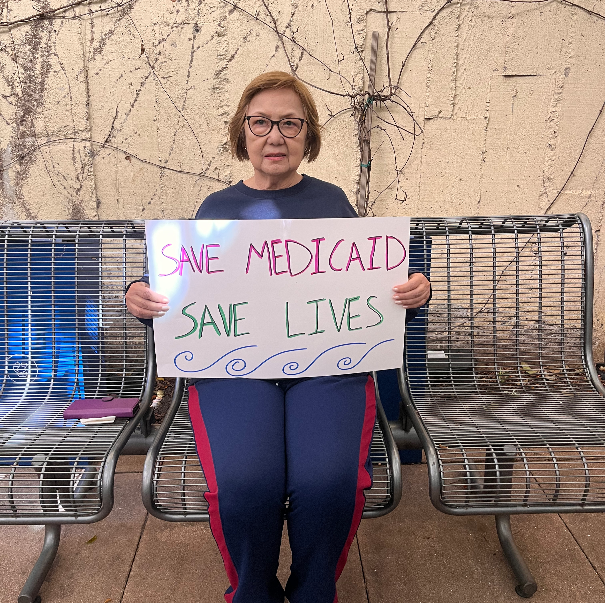 Una mujer filipina mayor está sentada en un banco, con una pared con ramas detrás de ella. Sostiene un letrero que dice: «Salven a Medicaid, salven vidas».