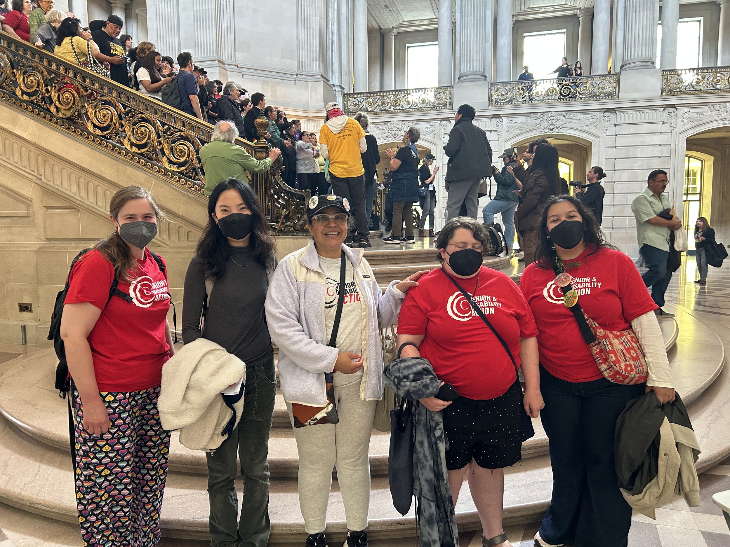 A group of five people, most of them masked and three in red SDA t-shirts, standing in front of the staircase at SF City Hall, with a crowd of people on the stairs behind them.