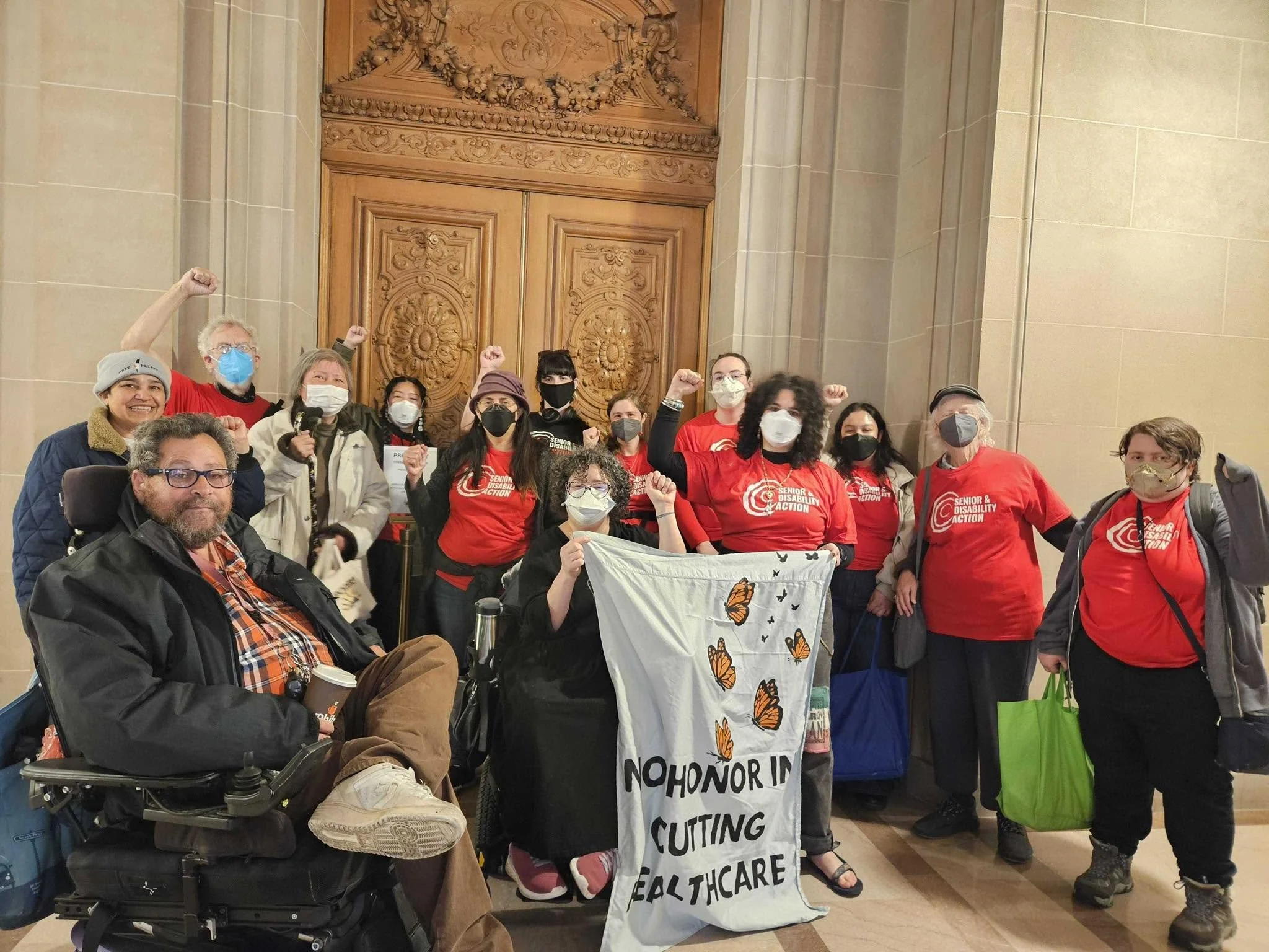 A group of around fourteen people in front of wooden doors in City Hall, some standing, and some in wheelchairs, most of them masked and wearing SDA t-shirts. In the front, they hold a banner that says "no honor in cutting healthcare."