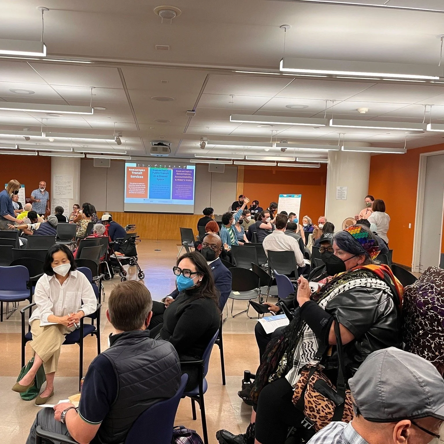 A large room with a screen at the front and groups of masked people sitting in circles on chairs and in wheelchairs