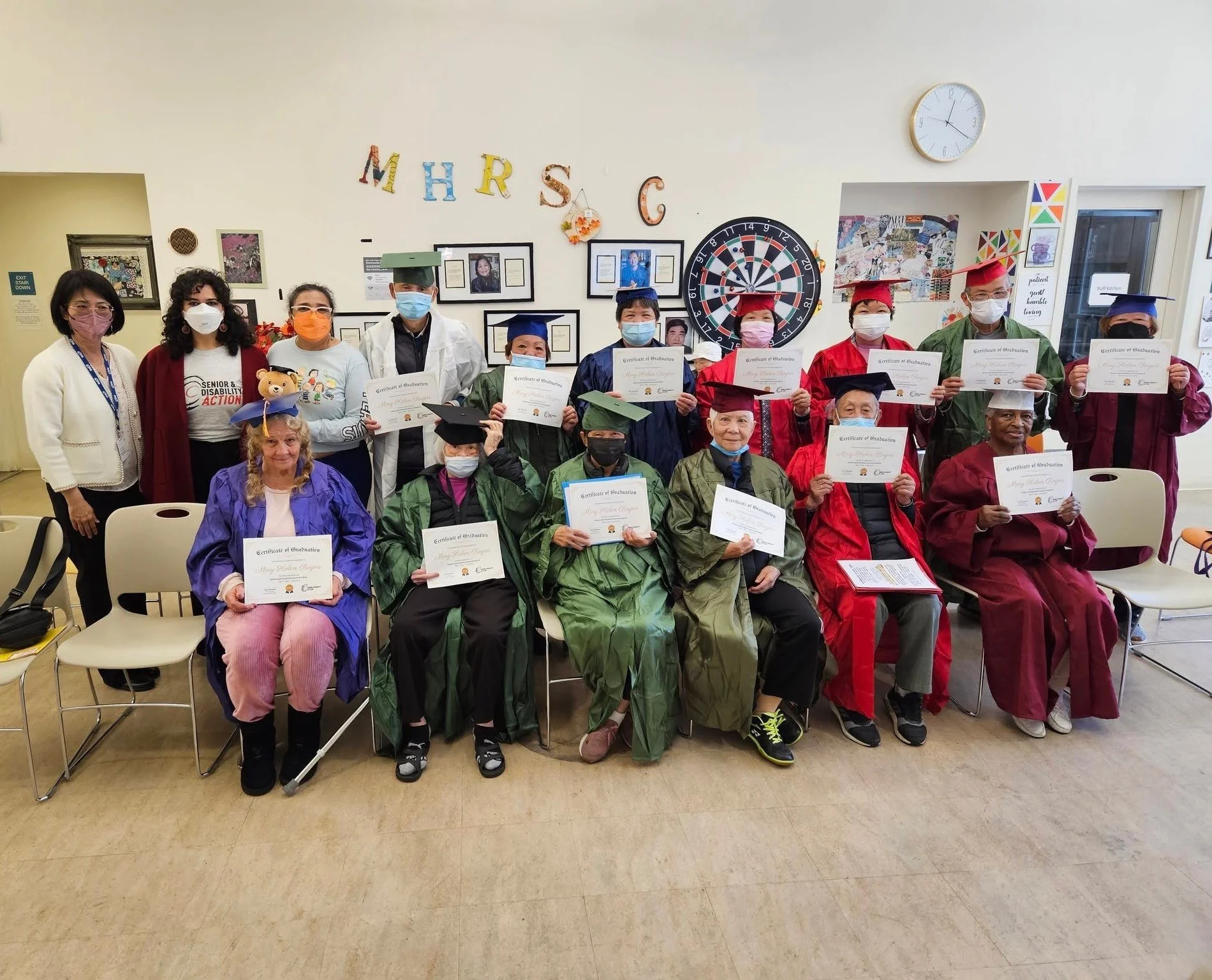 A group of masked people sitting and standing, many of them wearing graduation caps and gowns and holding certificates
