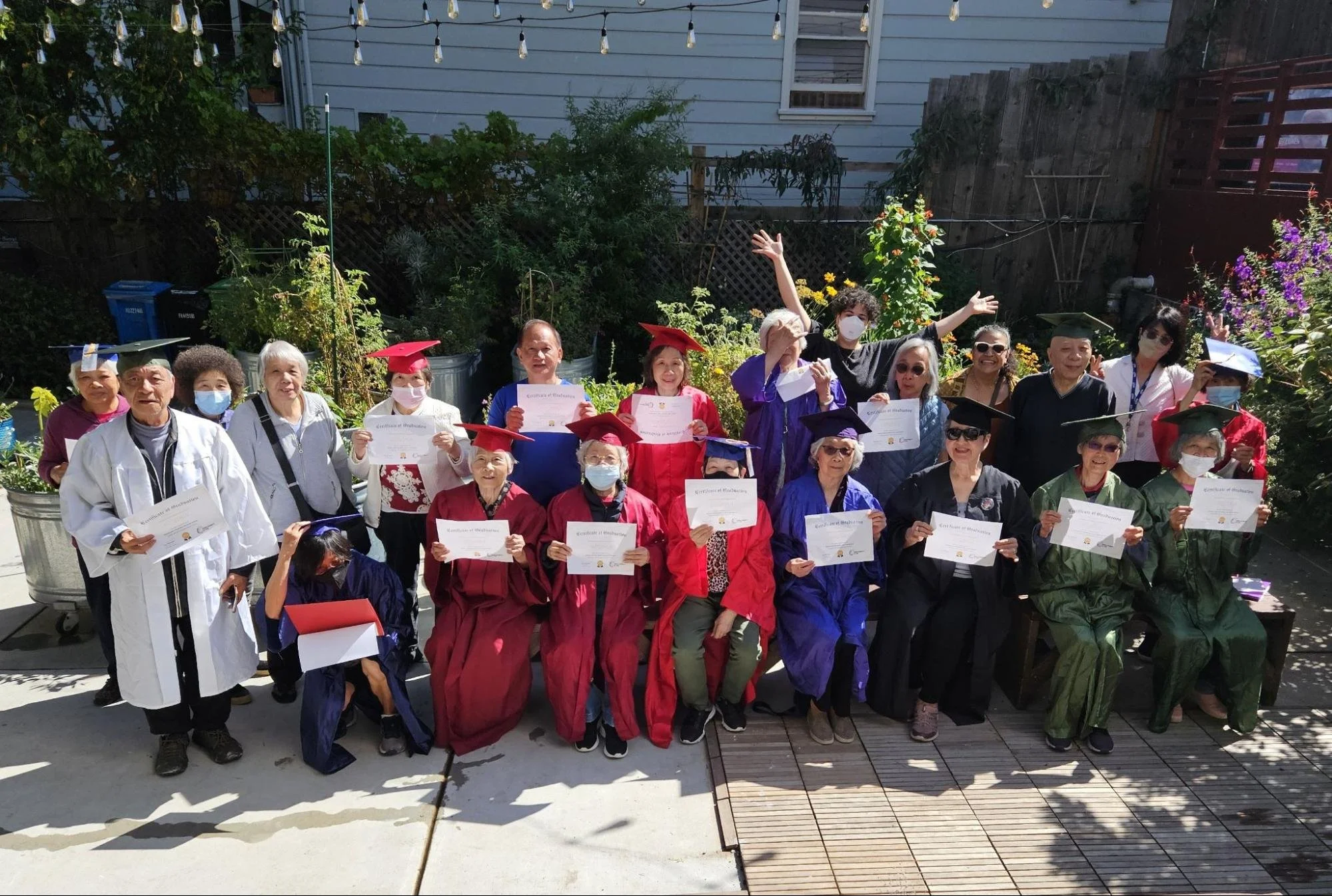 A group of around 15 seniors, many wearing graduation caps and gowns and holding diplomas, sitting and standing outside