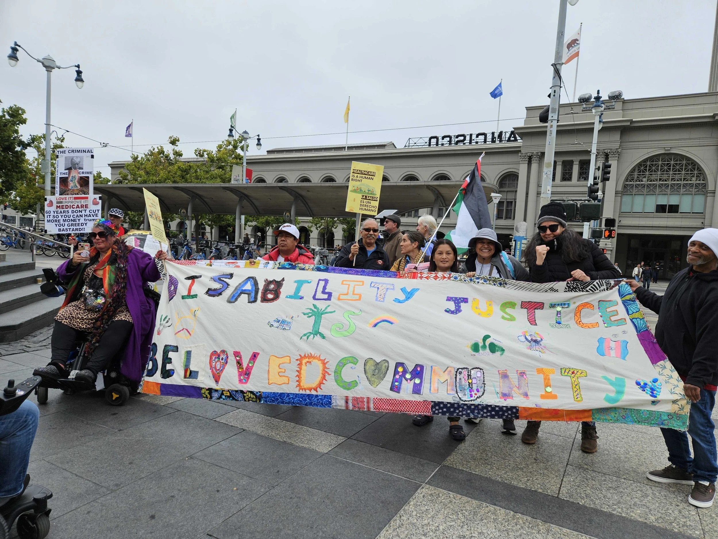A group of people outside in front of the SF Ferry Building holding a large fabric banner that says “disability justice is beloved community"
