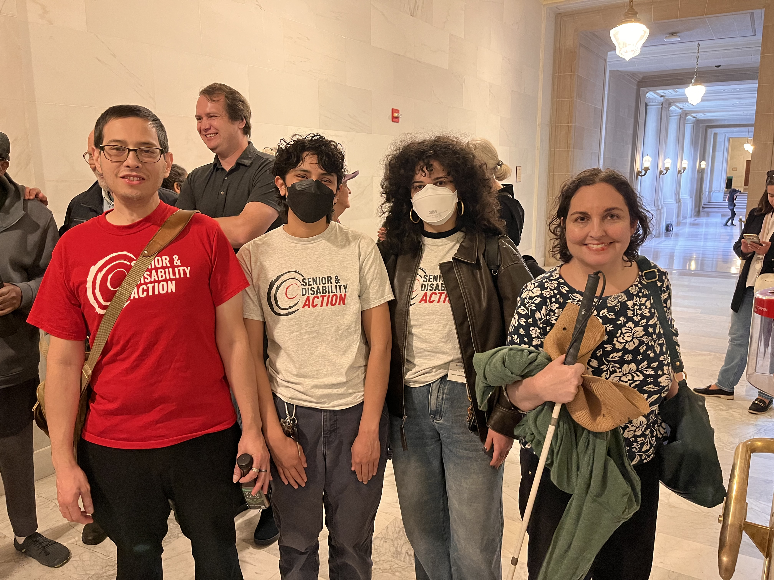 Four people standing in front of a white wall with a few other people in the background. Three of them are wearing Senior and Disability Action shirts, and one is holding a cane.