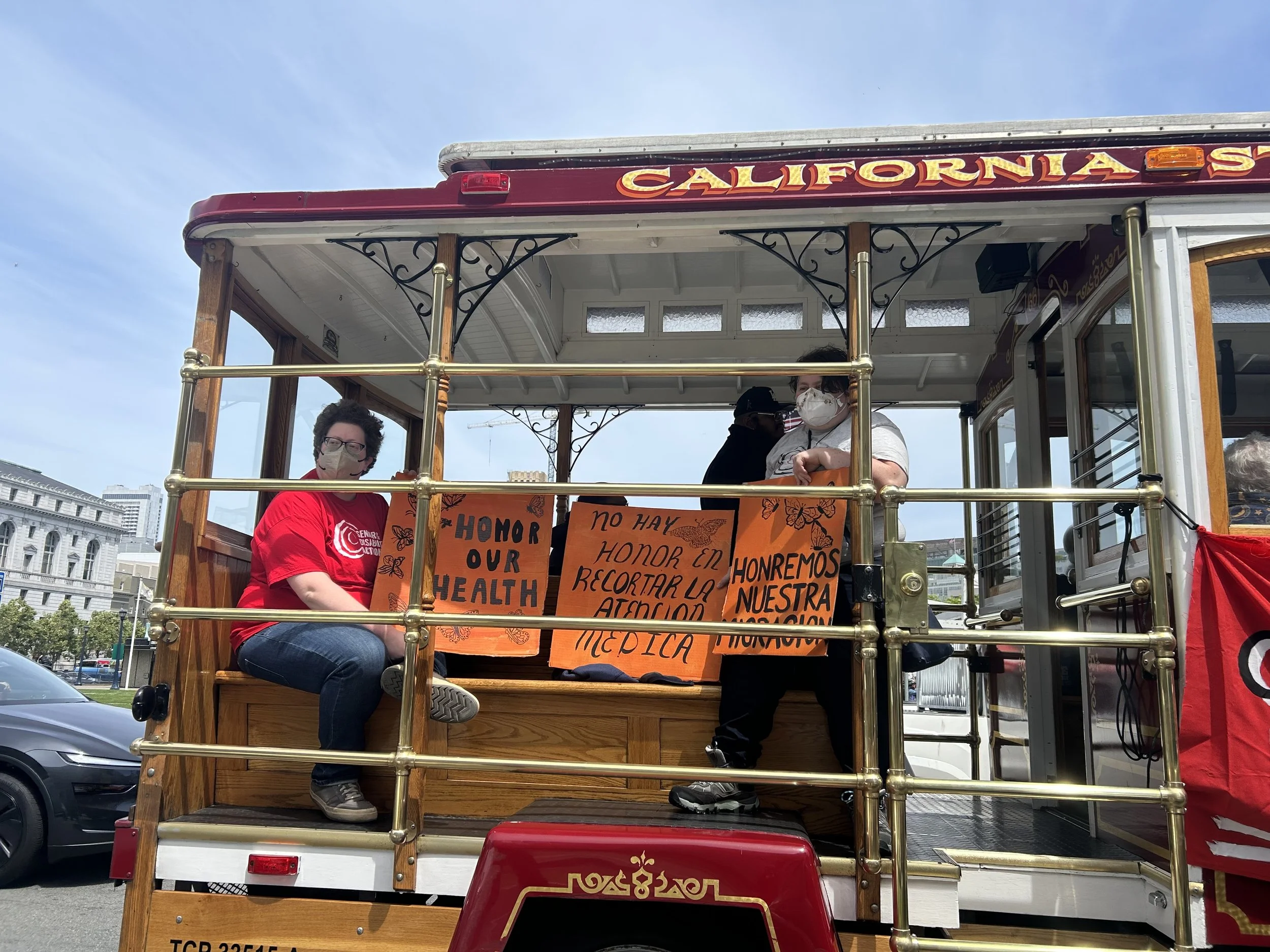 Two people on a cable car, holding signs that say "honor our health" and "no hay honor en recostar la atención médica."