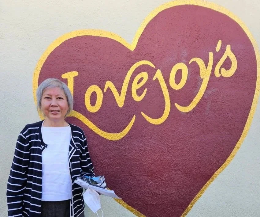A woman with short gray hair stands in front of a sign in the shape of a heart that says Lovejoy's