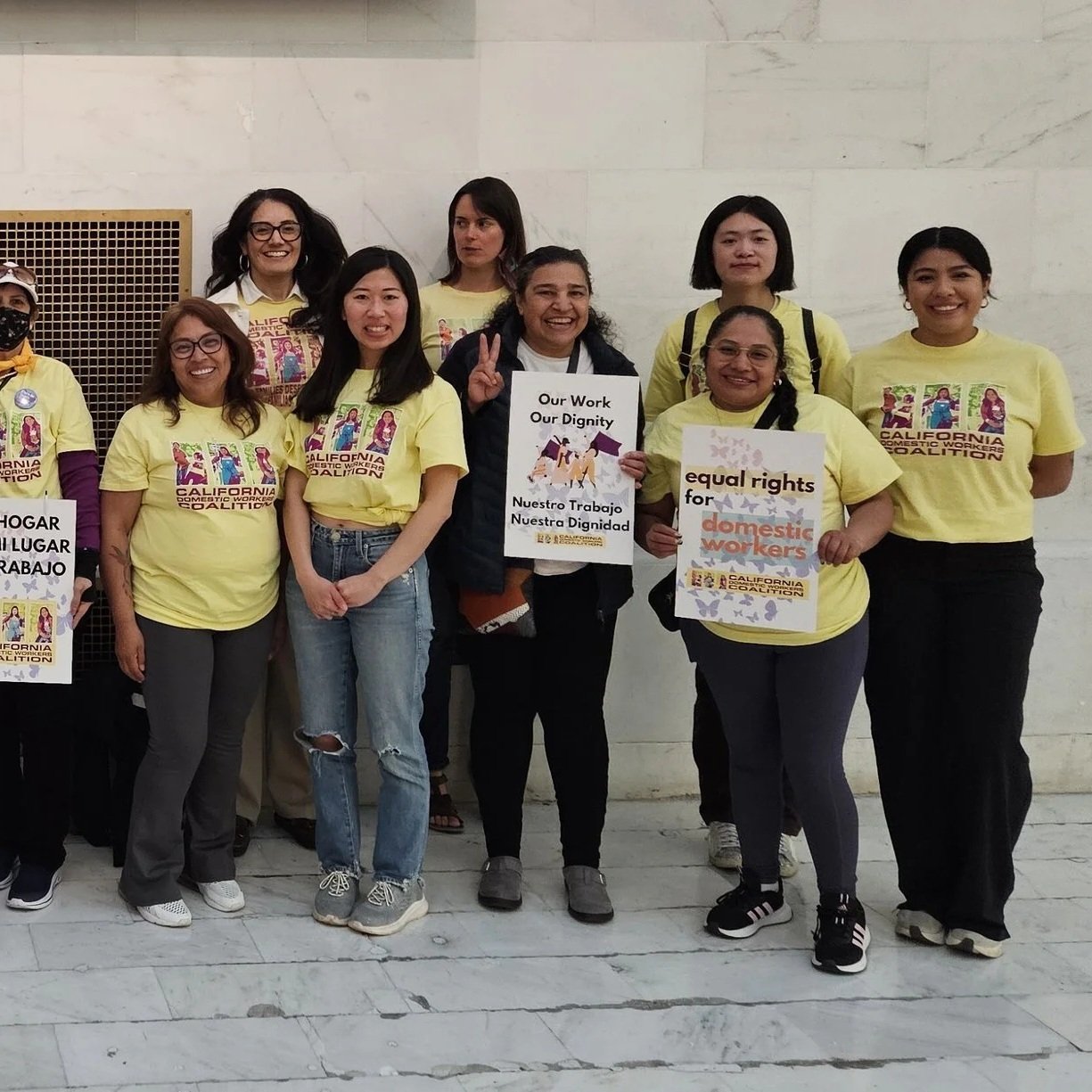 A group of people standing, many of them holding signs that say "Domestic workers rise up" and "Rise up for immigrant rights" and wearing t-shirts for California Domestic Workers Coalition