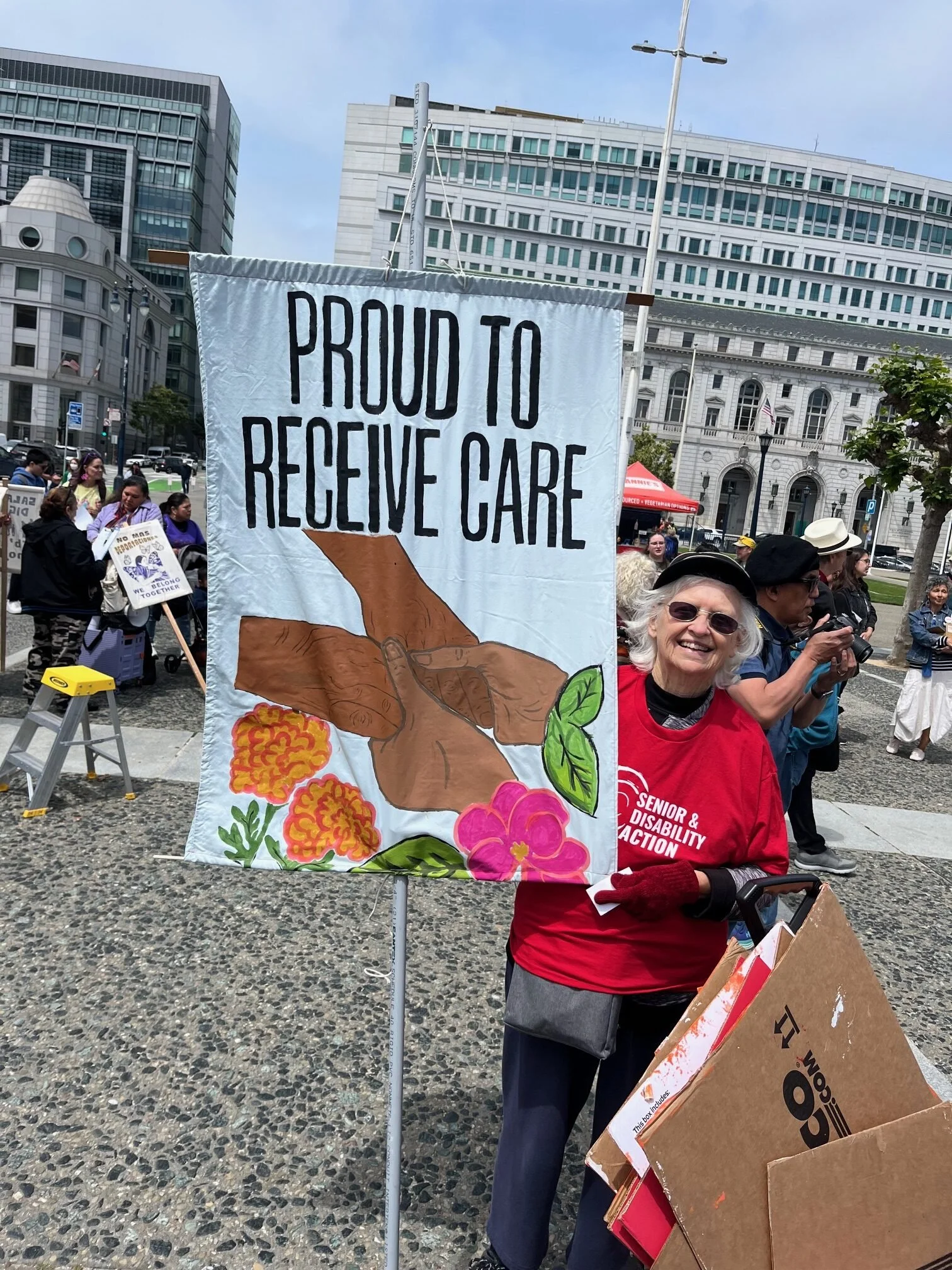 A smiling woman with white hair, a hat, and sunglasses holding a sign that says "proud to receive care" with an illustration of two pairs of brown hands holding each other and flowers at the bottom.