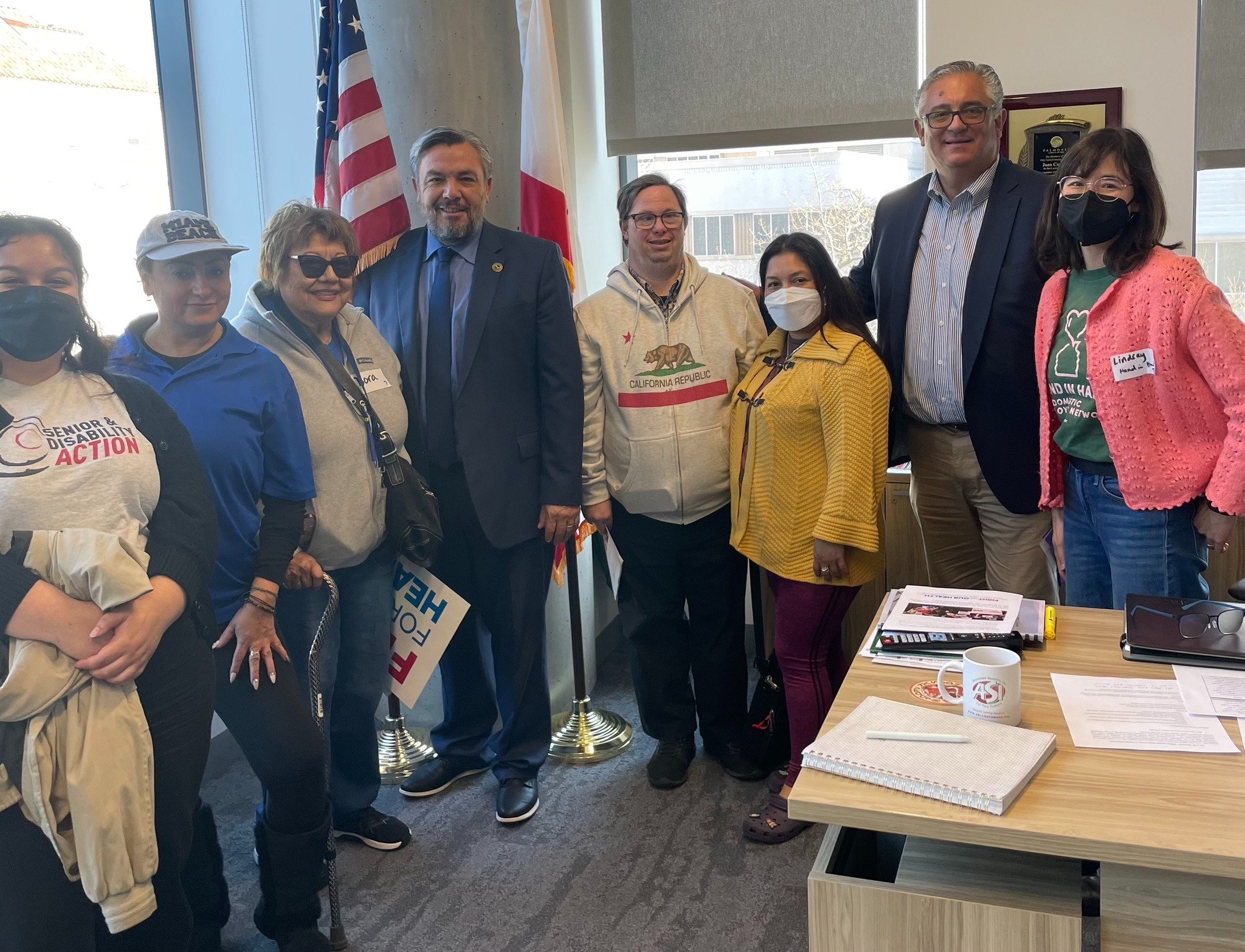 A multiracial group of seniors and disabled people standing in an office at the Capitol with two men in suits. They are standing behind a desk with notebooks on it, and some are wearing Senior and Disability Action and Hand in Hand shirts.