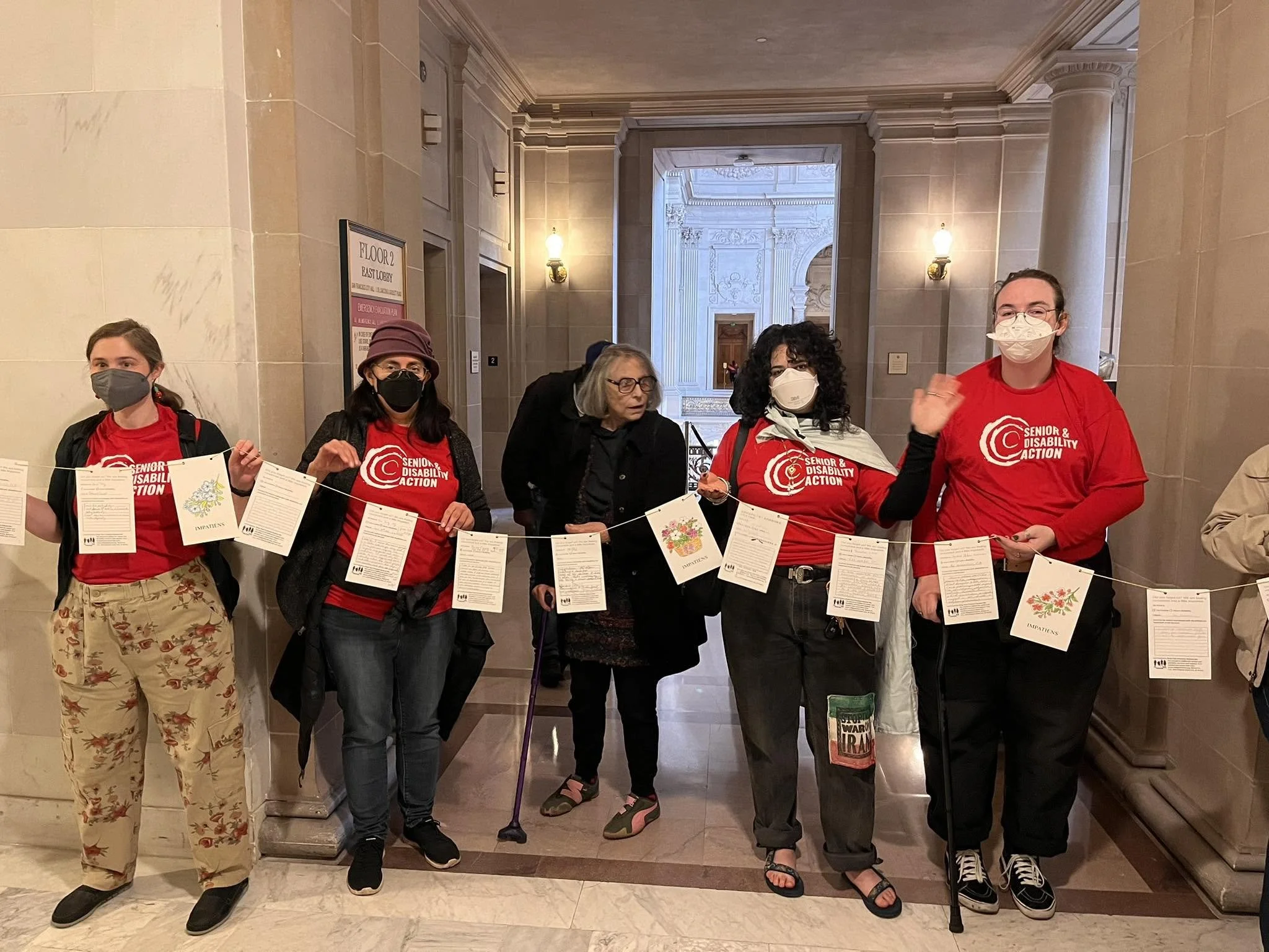A group of five people, most of them masked and wearing SDA t-shirts, holding a string of postcards (not legible in photo). They are standing in City Hall, with grey stone walls behind them.