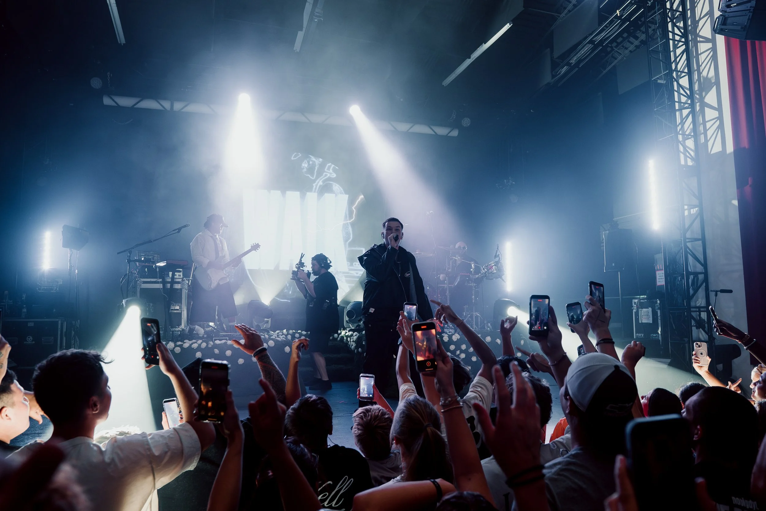 Live band performing on stage with audience watching in a dimly lit concert venue, with colorful stage lights and a dark curtain backdrop.