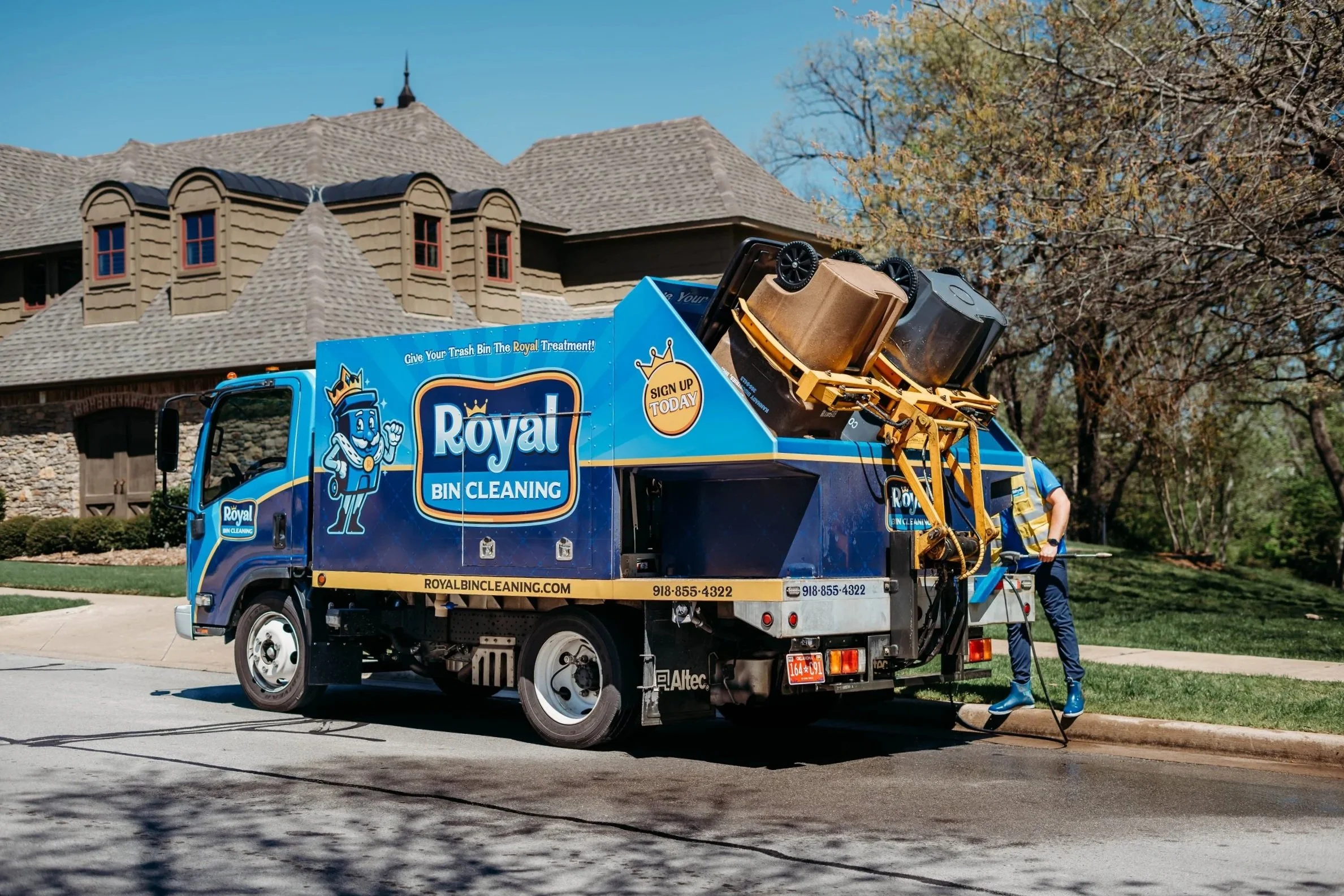 A blue Royal bin cleaning truck parked on a suburban street with a house and trees in the background. A worker is using equipment to clean the trash bins.