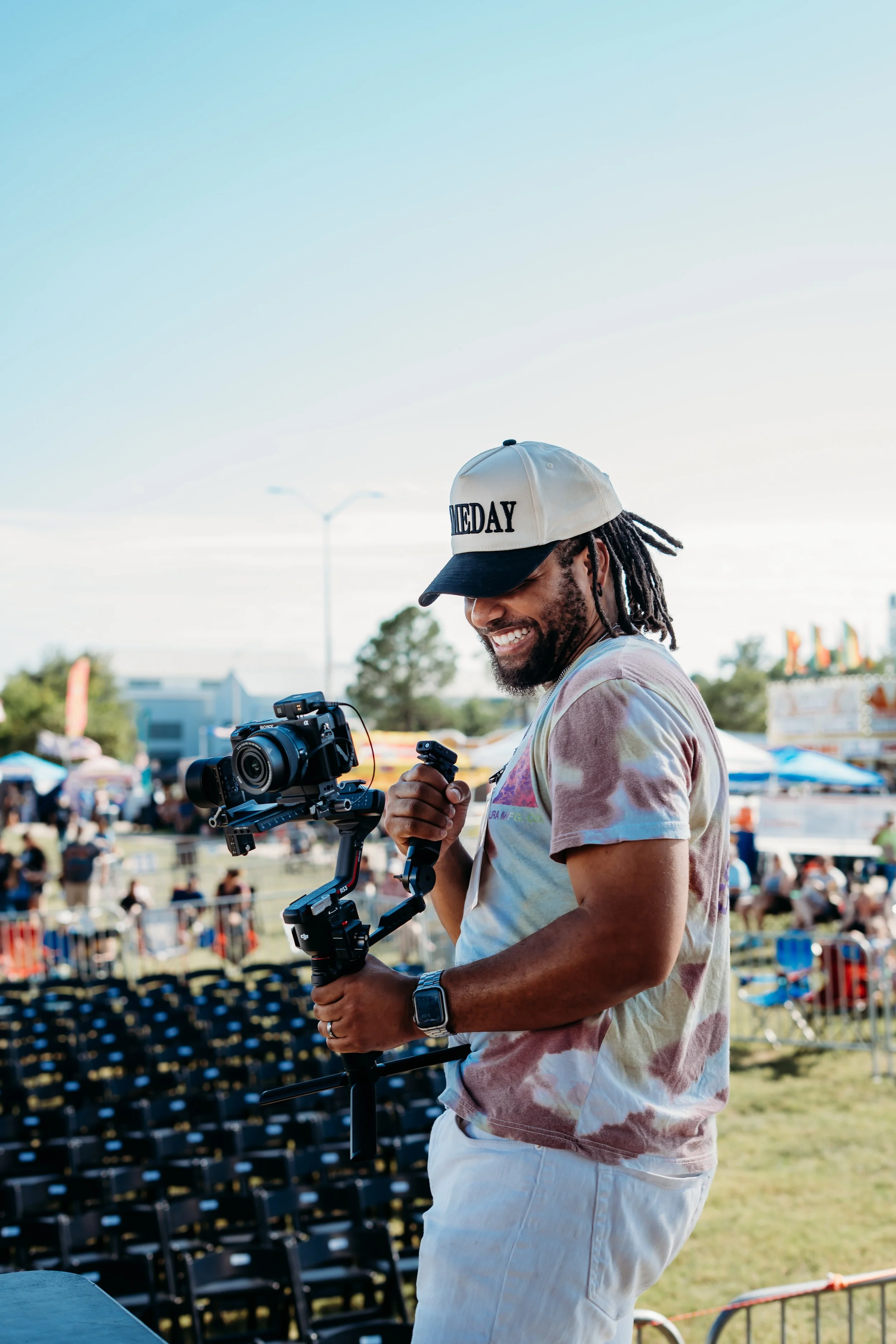 A man with dreadlocks wearing a cap and a tie-dye t-shirt is smiling and operating camera equipment outdoors at a festival or event, with a crowd and tents in the background under a clear sky.