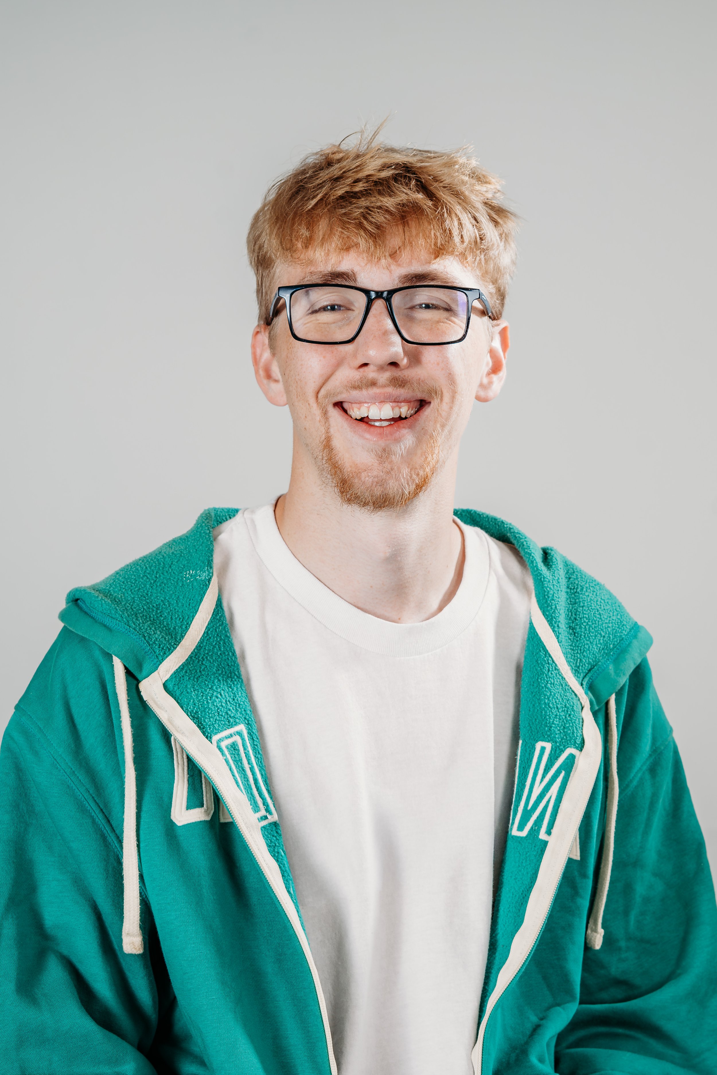 A young man with light skin, red hair, and glasses is smiling in front of a plain gray background. He is wearing a white t-shirt and a teal hoodie with white accents.