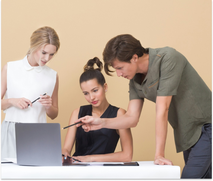 Three people surround a laptop that has been placed on a desk. A man is pointing to the screen explaining something to the woman sitting down in a chair. Another woman is on the left listening.